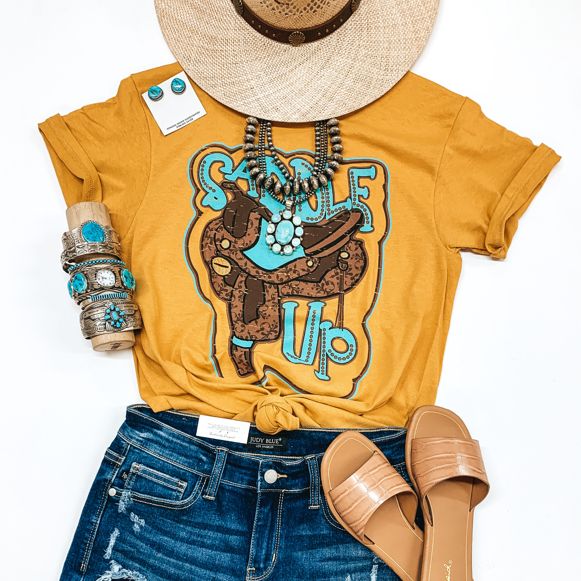 A mustard yellow graphic tee that says "Saddle Up". Pictured with genuine turquoise jewelry, a straw hat, denim shorts, and tan sandals.