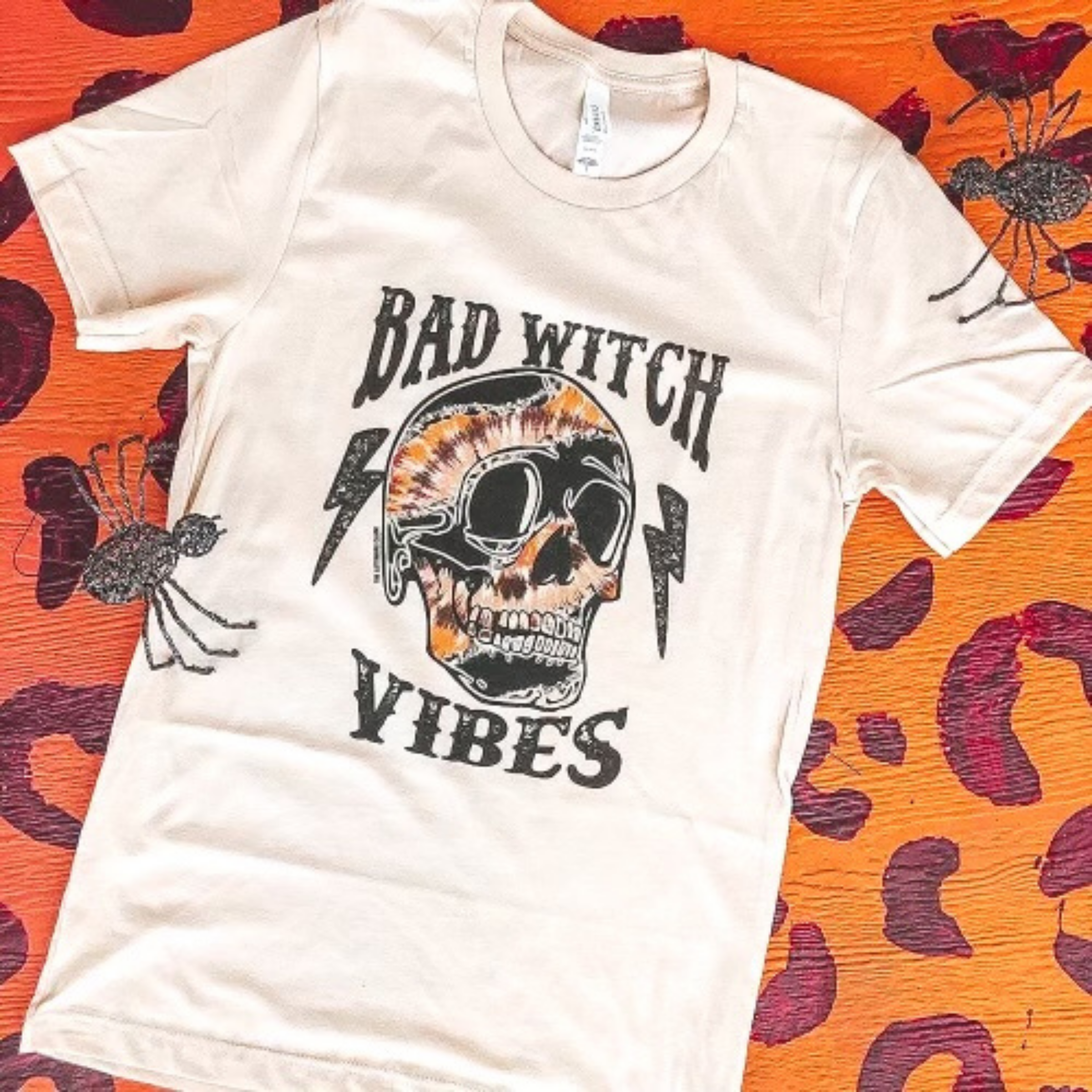 white graphic tee that reads "bad witch vibes" in black font with a multi colored skull in the center and lightning bolts. Laying on a leopard print backdrop with fake spiders as props. 