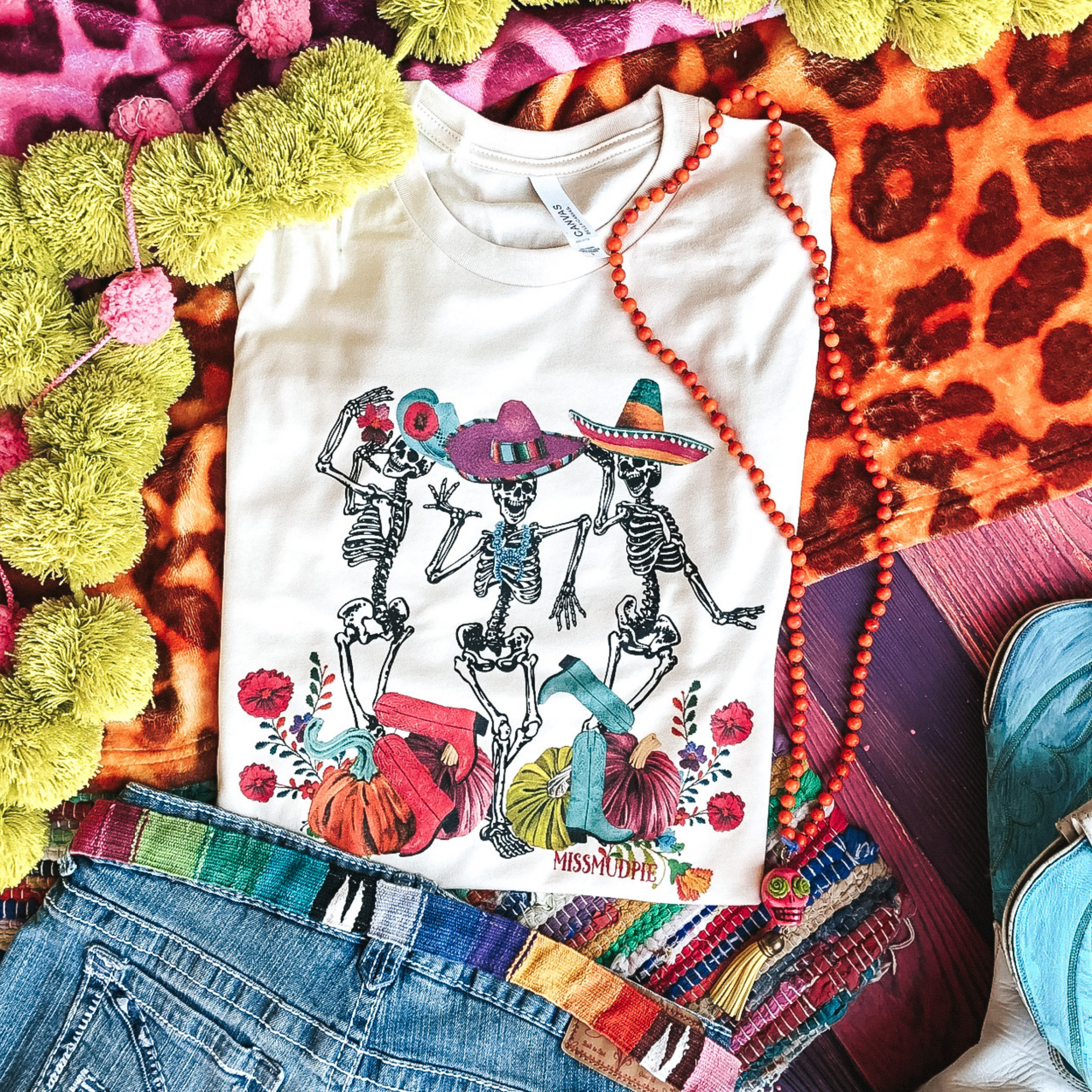 This is a cream shirt with 3 mariachi skeletons. Two out of the three skeletons are wearing sombreros and the other skeleton is wearing a hat with a flower on it. Two skeletons also have a pair of pink cowgirl boots and a pair of blue cowgirl boots. At the bottom of the shirt there are also colorful pumpkins and flowers.   