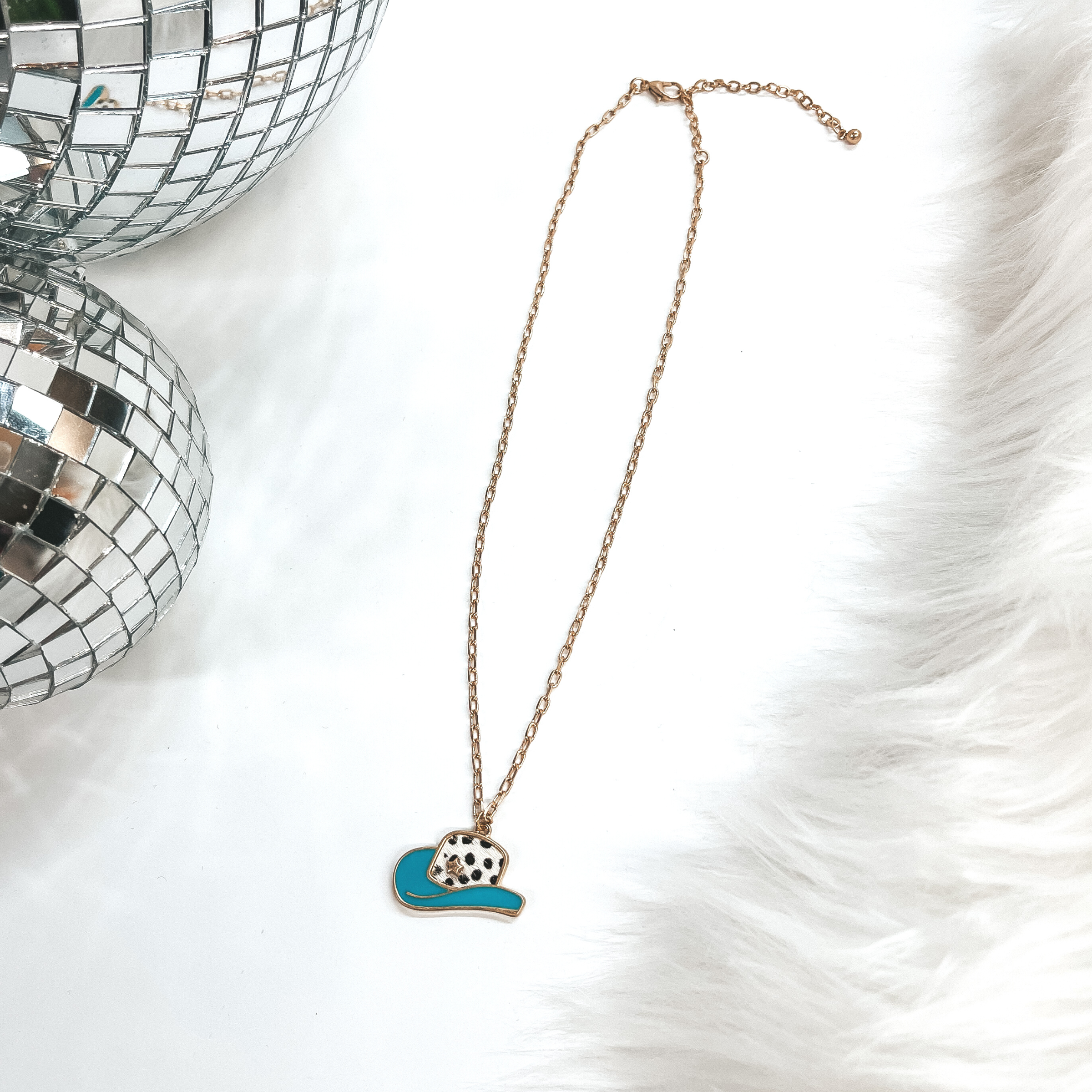This is a gold chain necklace with a hat pendant in  a gold setting. The hat pendant is turquoise and has  a dotted print with a gold star. This necklace is  taken on laying on a white background with a white  fur carpet in the side and two disco ball as decor.