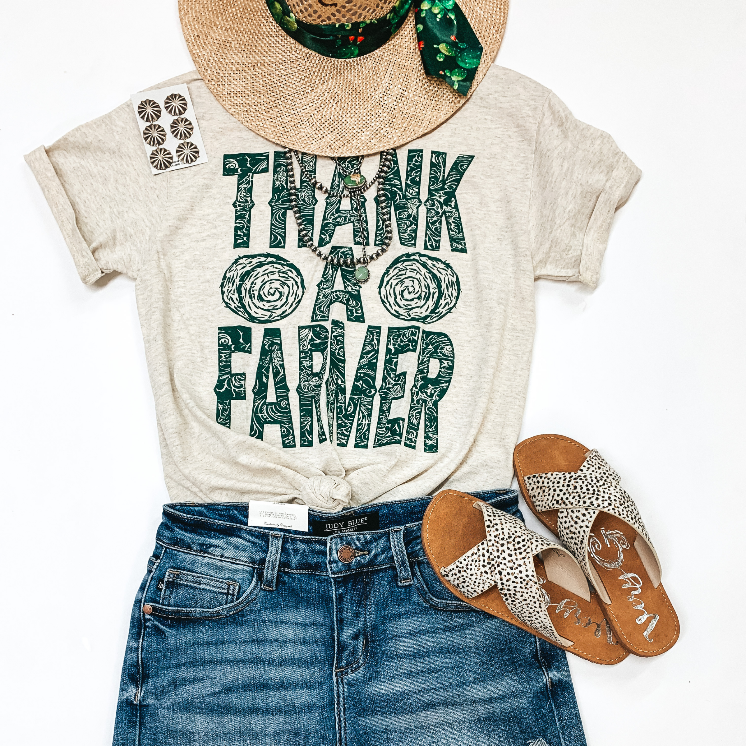 With a front knot, a light tan shirt with "Thank A Farmer" written in the middle. in green. 2 haybales are on either side of the shirt, also in green.  Below the shirt is a pair of blue jean shorts with black and white cross sandals beside them. A Charlie 1 Horse Straw Hat is on the top side of the shirt and a pair of concho earrings lays alongside the left of the shirt. Picture is on a white background.  2 navajo necklaces lay along the neckline of the shirt. 