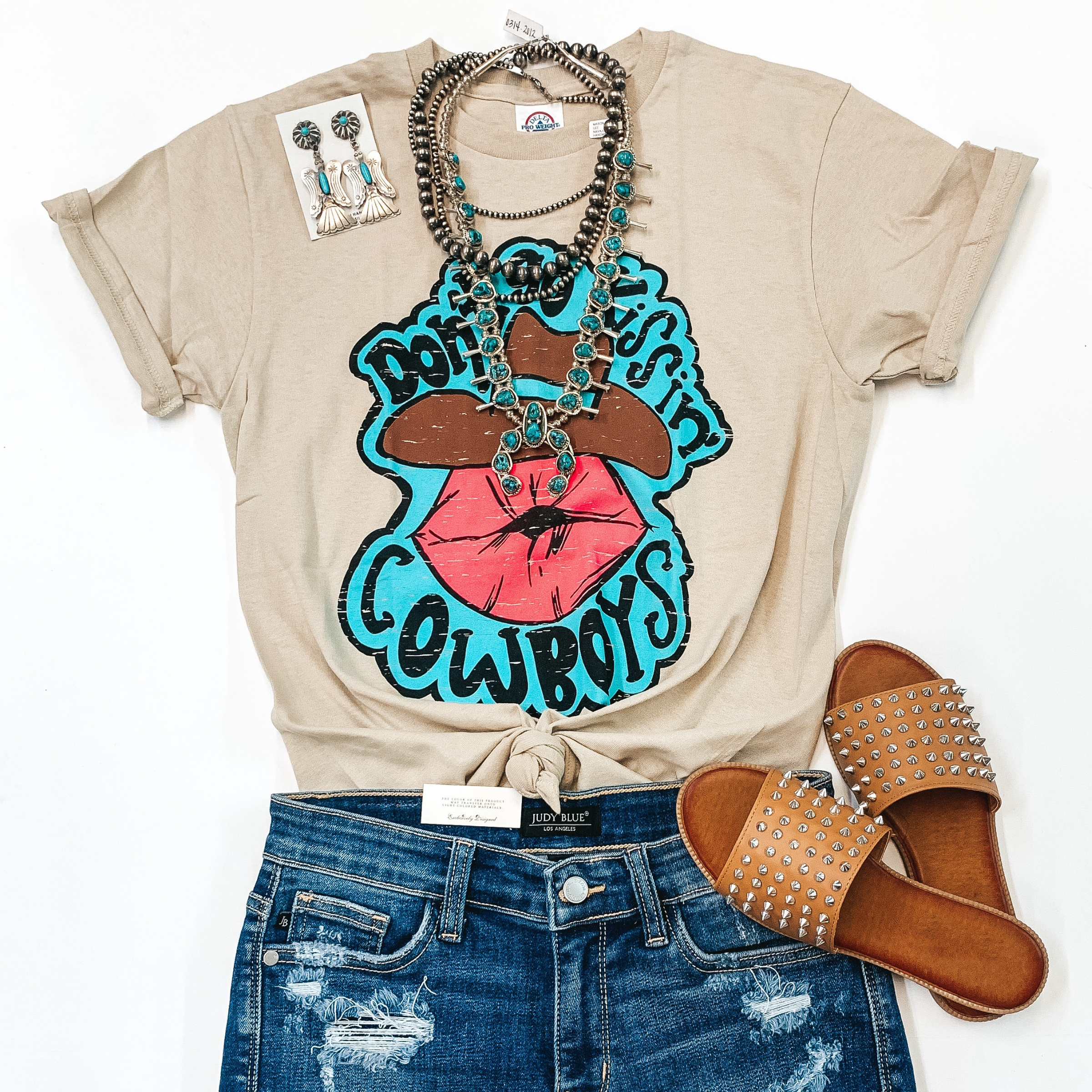 A beige tee shirt with a graphic that says " Don't Go Kissing Cowboys." Pictured with denim shorts, tan sandals, and genuine Navajo jewelry.