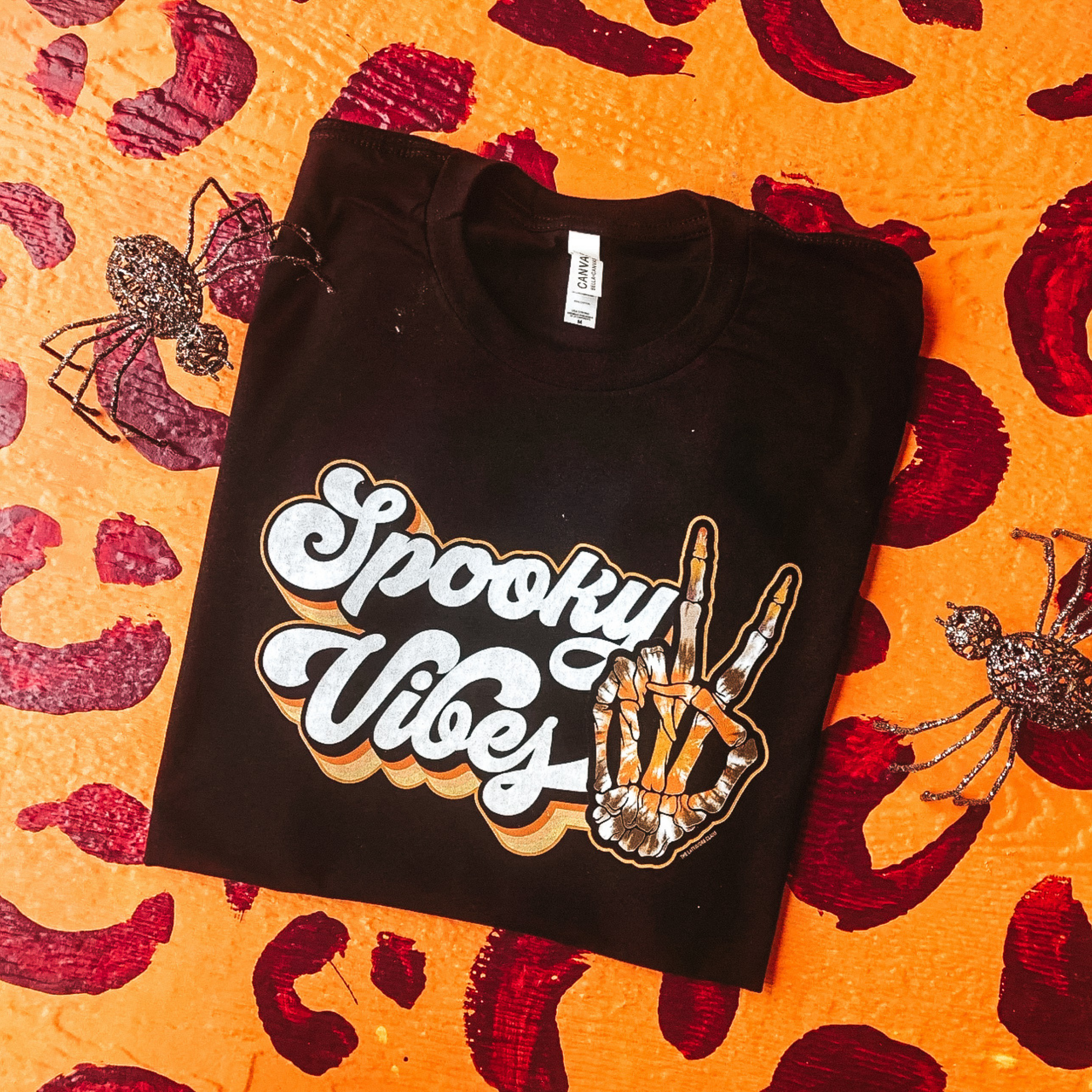 black graphic tee that reads "spooky vibes" in a groovy font with a skeleton hand doing a peace sign hand gesture. Tee is laying on a leopard print backdrop with fake spiders as props. 
