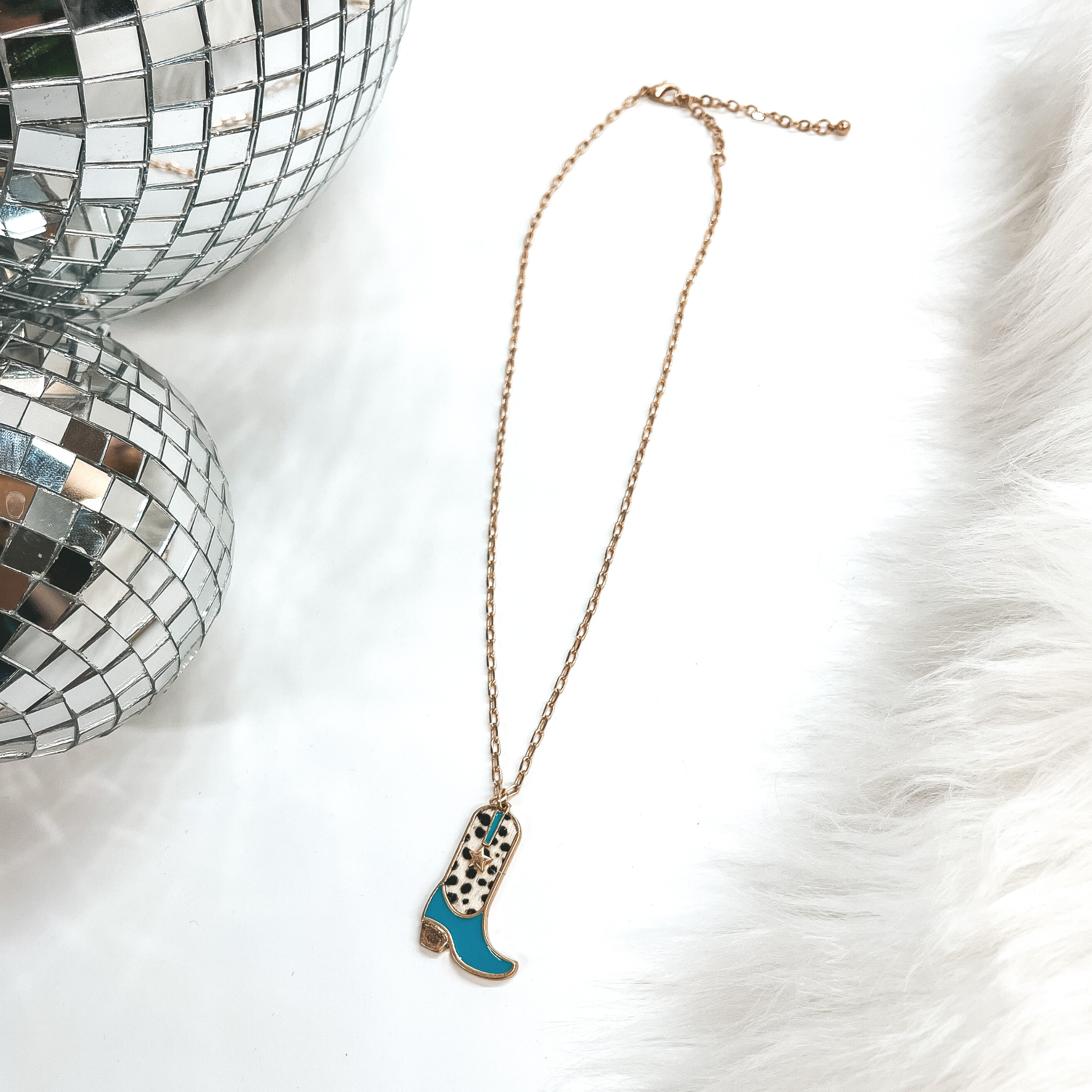 This is a gold chain necklace with a boot pendant  in a gold setting. The boot pendant is turquoise and  has a dotted print with a gold star. This necklace  is taken on laying on a white background with a  white fur carpet in the side and two disco ball as  decor.