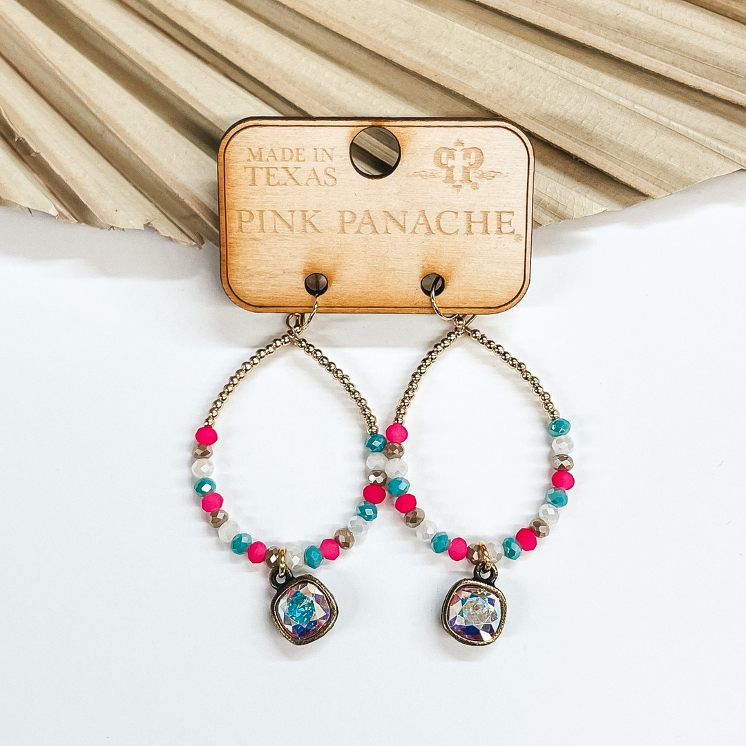 Fish hook gold teardrops with multicolored beads  and AB cushion cut pendant. The teardrop has small  gold beads on the top half. The bottom half of the teardrop have beads in light beige, white, light blue, and pink. Taken on a white  background and leaned up against a dried up palm leaf.