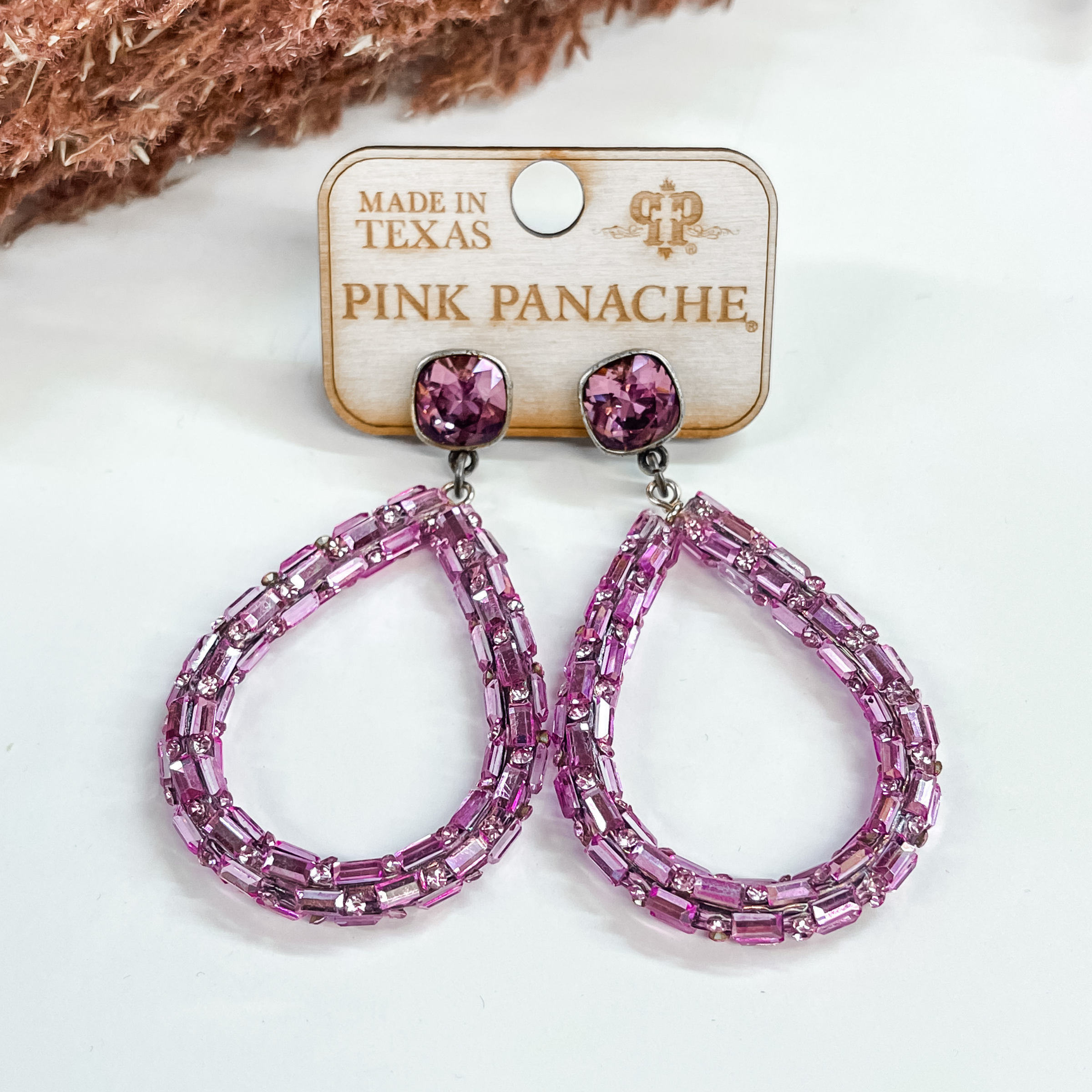 Purple cushion cut crystal studs with hanging  teardrop dangle in purple.These earrings  are pictured on a white background with a dark brown plant in the back as decor.