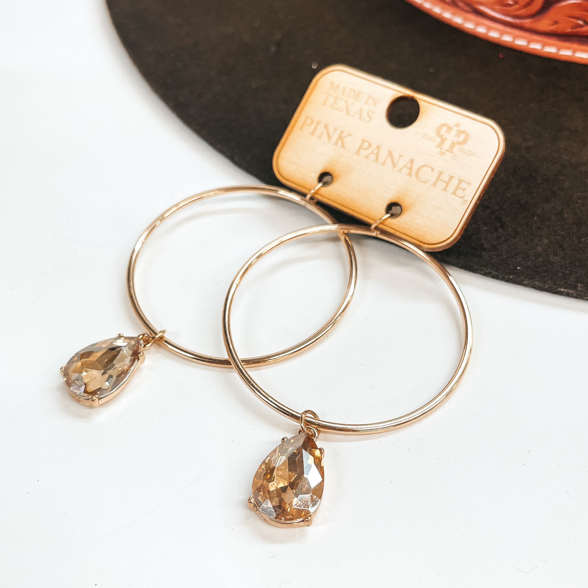Fish hook gold hoop earrings with a light silk  teardrop shaped crystal pendant in the center.  Taken on a white  and leaning against a dark brown hat.