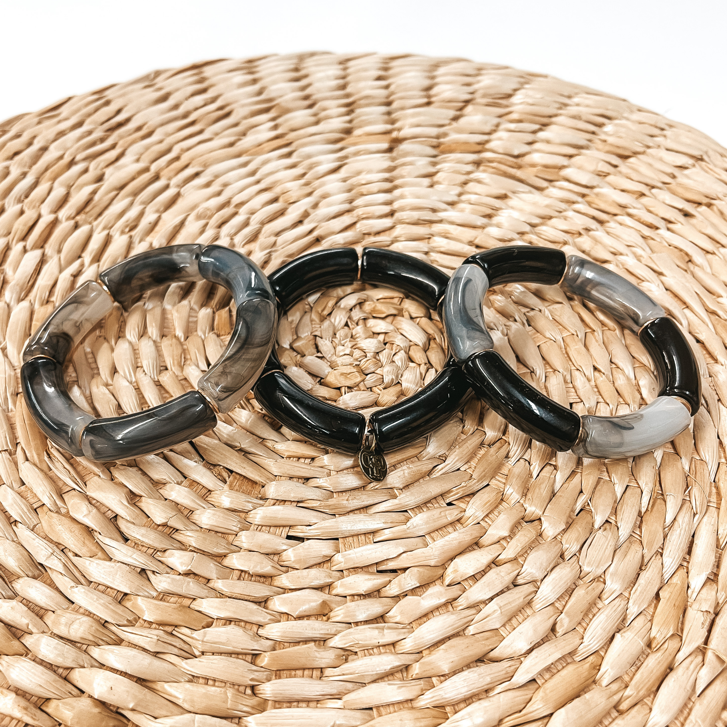 These are three acrylic tube bracelets in black  with different acrylic patterns and gold spacers.  The bracelet in the  left is a clear grey and black marble tubes. The middle one is all solid black.  The right one has a mix of solid black and marble  tubes. Taken on top of a bamboo stool and white background.
