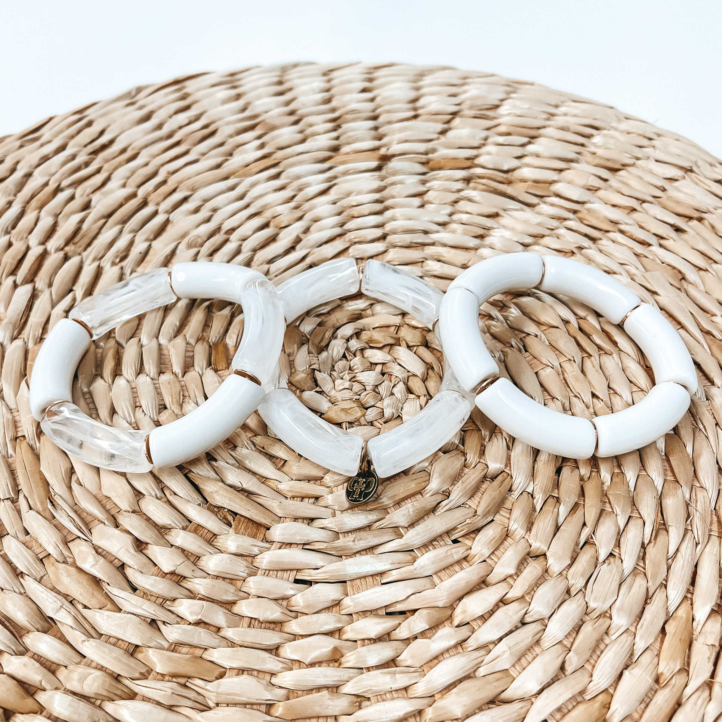 These are three acrylic tube bracelets in white  with different acrylic patterns and gold spacers. The bracelet in the  left is a mix of solid white and marble tubes. The middle one is a mix of clear and white marble tubes.  The right one is all solid white tubes. Taken on top of a bamboo stool and white background.