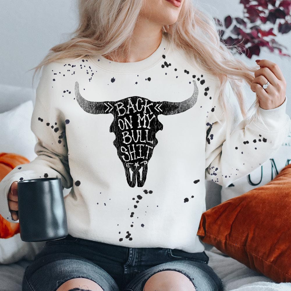 This white sweatshirt includes a crew neckline, long sleeves, and cute hand drawn design of a black bullhead with the words "Back On My Bull Shit" in white featured inside. Black paint splatters are all over the sweatshirt as well. 
