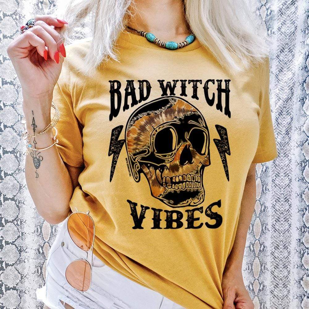 Model is wearing a yellow graphic tee that reads "bad witch vibes" in black font and a multi colored skull and lightning bolts.  Model is in front of a snakeskin printed background and has it paired with white shorts.