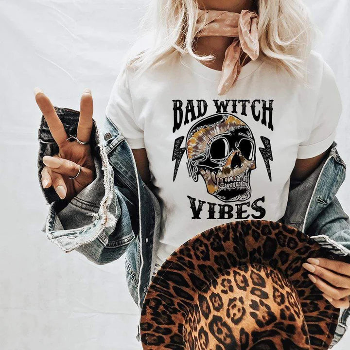 Model is wearing a white graphic tee that reads "bad witch vibes" in black font with multi colored skull and lightning bolts. Model has it paired with a denim jacket, pink wild rag, and a leopard print hat. 