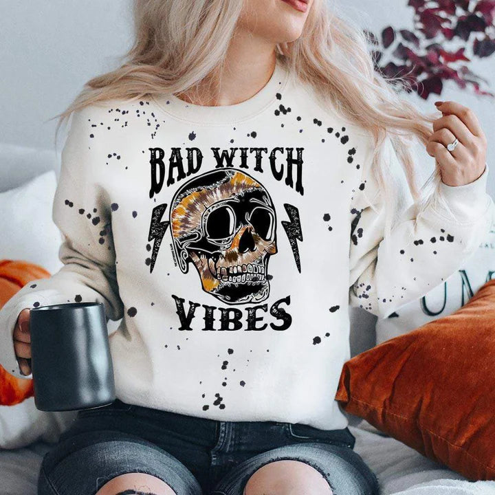 model is wearing a white sweatshirt which reads "bad witch vibes" in black font with a multicolored skull and lightning bolts. Model is sitting in front of a fall background and has it paired with black, ripped jeans.  