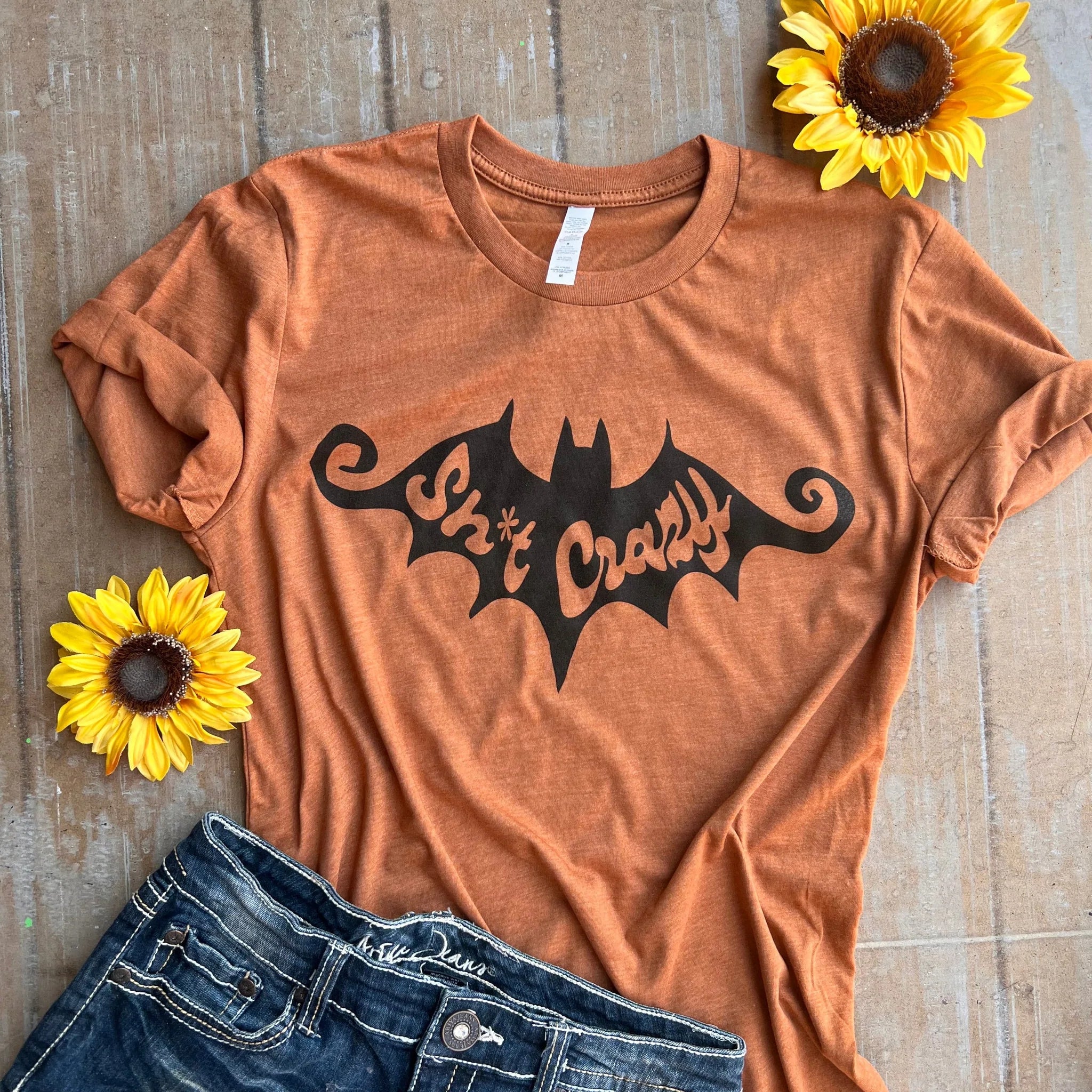 This is a short sleeve shirt in harvest orange with a bat with the words sh*t crazy in the middle of the bat. There are two sunflowers around the shirt with a pair of blue jeans at the bottom of the picture.