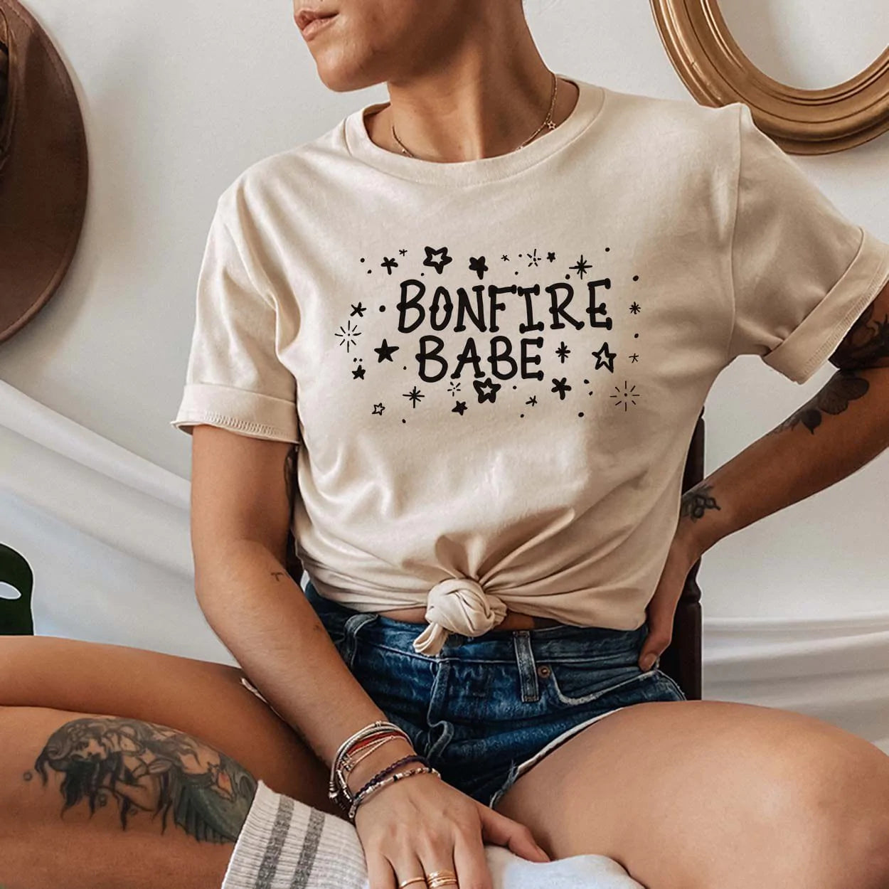 Model wearing cream t-shirt with the words bonfire babe and star around the words in black. You will also find decorative fixtures in the background. 