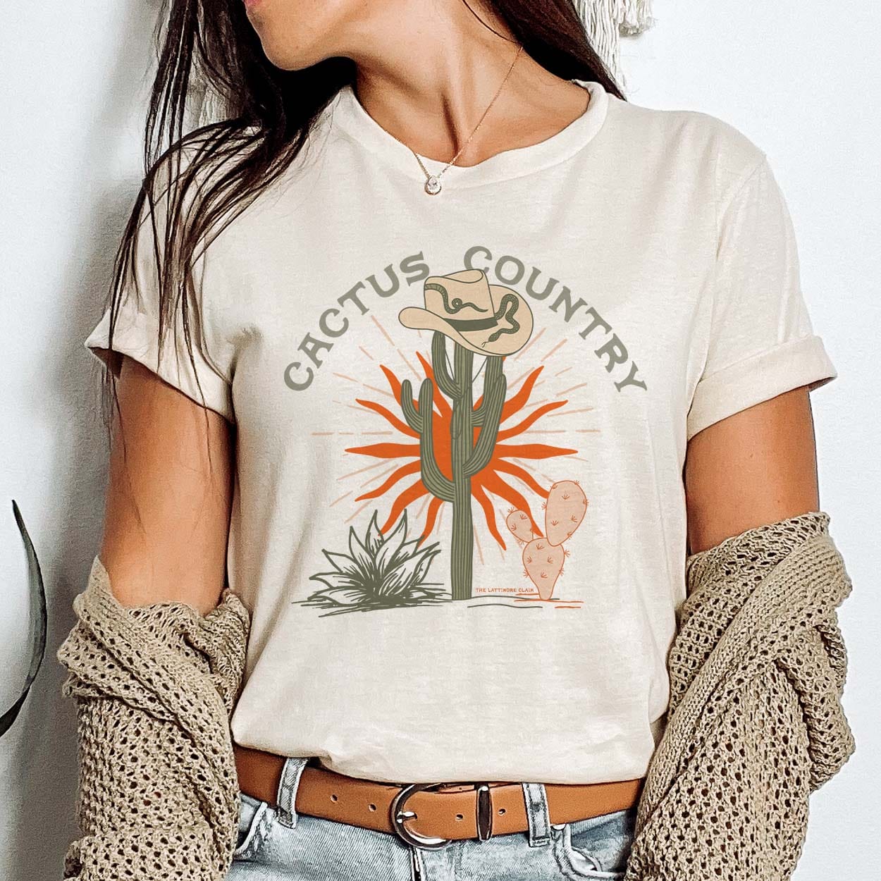 A cream colored short sleeve tee with cuffed sleeves, a graphic of a cactus wearing a tan cowboy hat with a snake, a little orange cactus, green foliage, an orange sun behind the cacti, and the words "cactus country" above the graphic. Item is pictured on a plain white background.