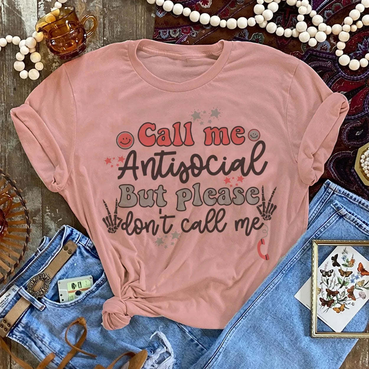 This desert rose pink graphic tee includes a crew neckline, short sleeves, and cute hand drawn design of skeleton hands holding up peace signs, stars, and smiley faces. The words "Call Me Antisocial But Please Don't Call Me" are stacked in the middle of the tee. This tee is shown in this photo as a flat lay, styled with belted denim jeans. 