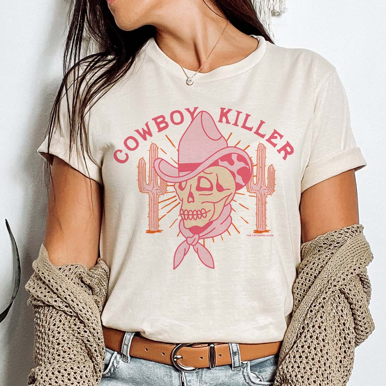 A cream colored short sleeve shirt with cuffed sleeves featuring a graphic of a pink skull with a cowboy hat and bandana and cacti on either side with rays coming out behind the skull. The text "cowboy killer" is in pink above the graphic. Item is pictured on a plain white background.