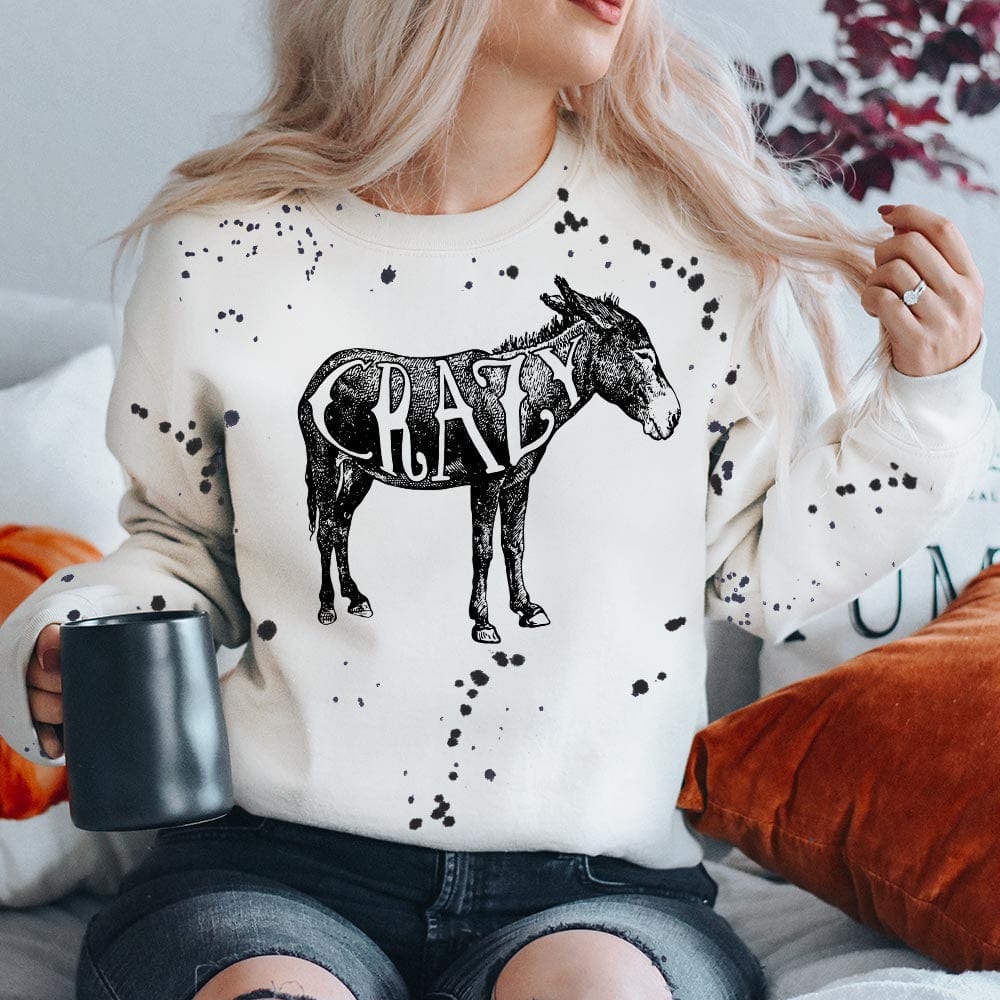 This white sweatshirt includes a crew neckline, long sleeves, and cute hand drawn design of a donkey with the word "Crazy" inside. There are also black paint splatters all over the sweatshirt. 