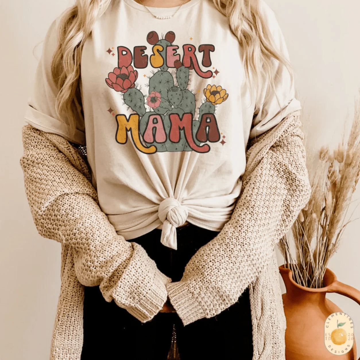 A cream colored short sleeve crew neck tee featuring a graphic of a cactus with pink and orange flowers and the text "desert mama" in coordinating colors with the flowers. Item is pictured on a plain white background