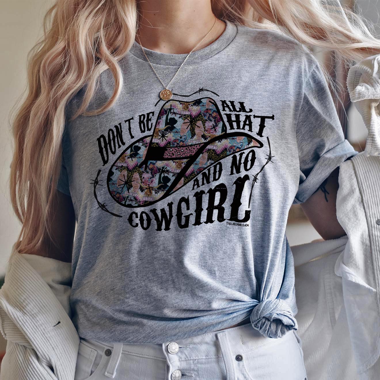 This heather grey Bella + Canvas graphic tee includes a crew neckline, short sleeves, and cute hand drawn design. of a cowboy hats when fun + bright prints, and the words "Don't Be All That And No Cowgirl". It also has black barbwire intertwined with the words and around the hat. This sweatshirt is shown in this photo to be styled with a necklace, white shacket, and light wash denim jeans. 