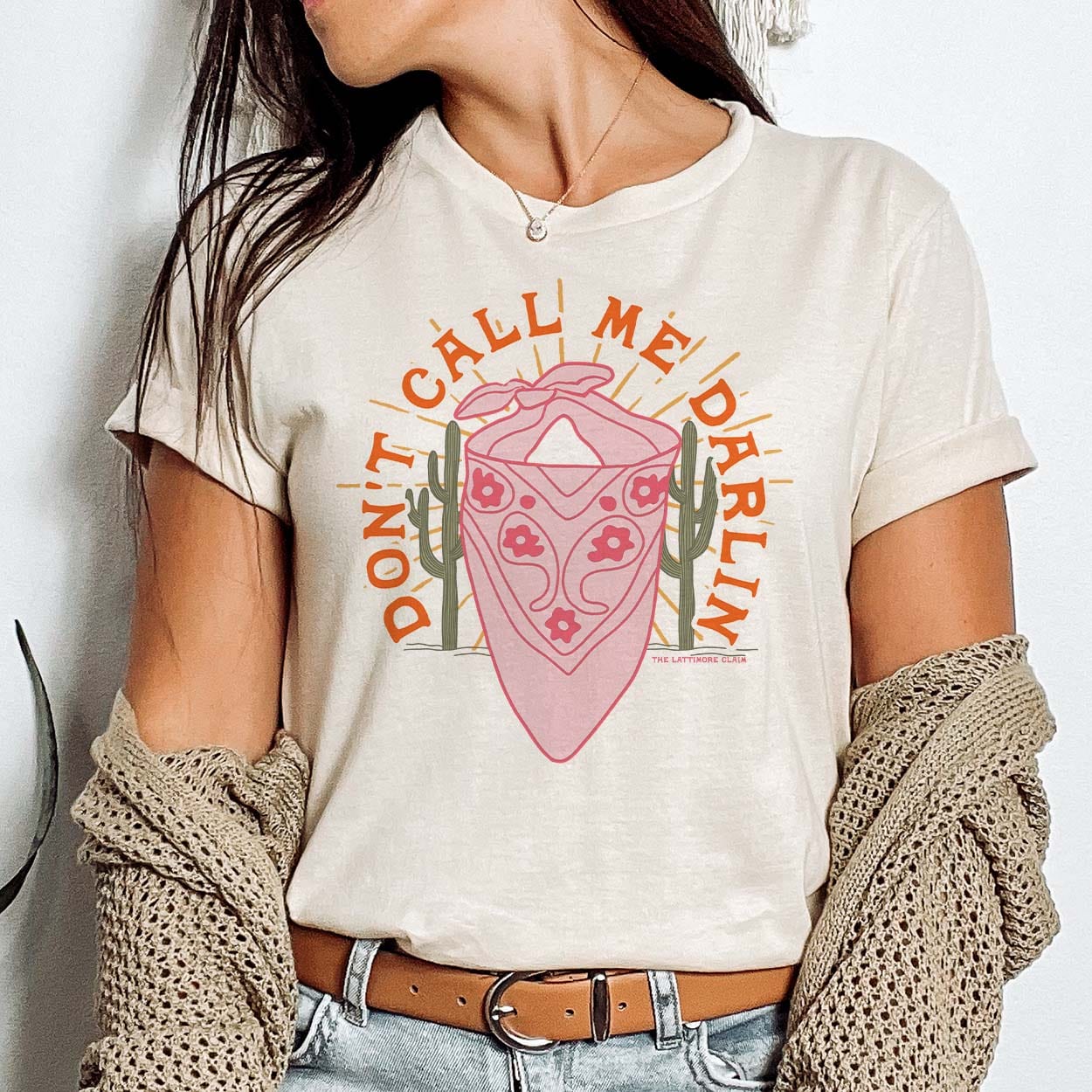 A cream colored short sleeve shirt with cuffed sleeves featuring a graphic of a pink floral bandana with a green cactus on both sides and light orange rays coming from behind the bandana. The text "Dont call me darlin" is curved around the top of the graphic in orange. Item is pictured on a white background