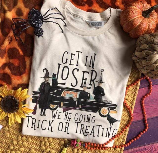 This is a cream shirt that says get in loser were going trick or treating. In the middle of the words there is a black hearse with 3 witches carrying brooms. Two of the witches are wearing black and white striped hats while the other one is wearing an all black hat. There is also a black crow and a black cat next to the hearse.