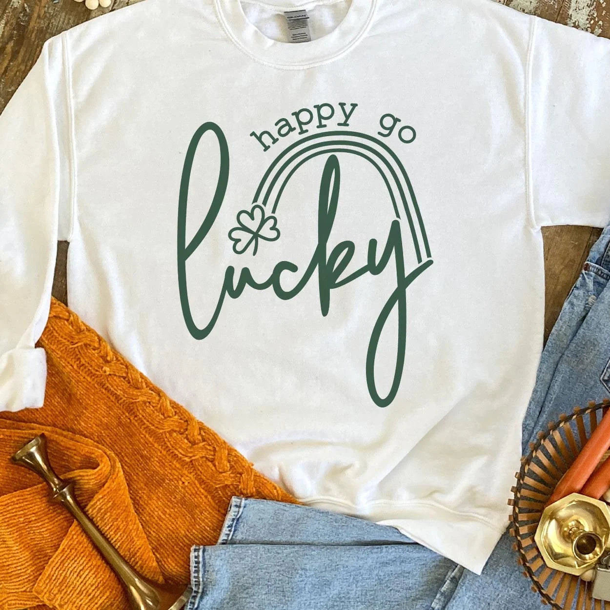 A white crew neck sweatshirt featuring the words "Happy go lucky". "Lucky" being in the center in cursive while "happy go" is above "lucky" in a smaller font. Between the two is a small rainbow with a clover at the end. Item is pictured on a brown background.