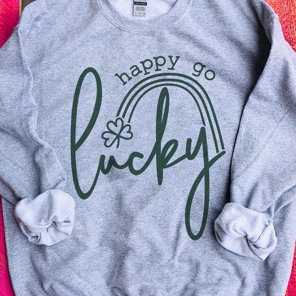 A gray crew neck sweatshirt featuring the words "Happy go lucky". "Lucky" being in the center in cursive while "happy go" is above "lucky" in a smaller font. Between the two is a small rainbow with a clover at the end. Item is pictured on a pink background.