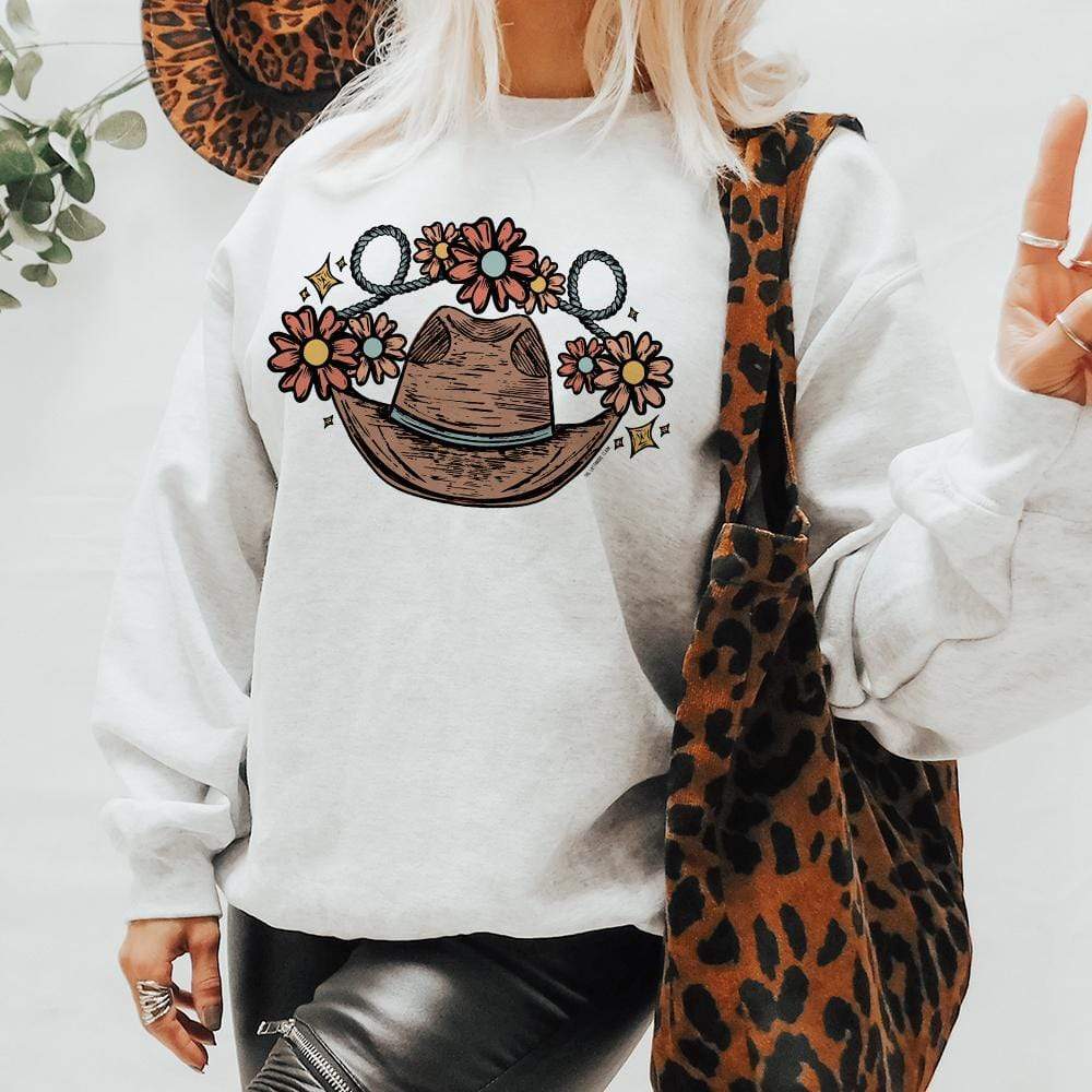 This white sweatshirt includes a crew neckline, long sleeves, and cute hand drawn design of a brown cowboy hat with a turquoise hat band, rope, and flowers. It is shown in this picture styled with faux black leather leggings, silver jewelry, and a leopard print purse. 