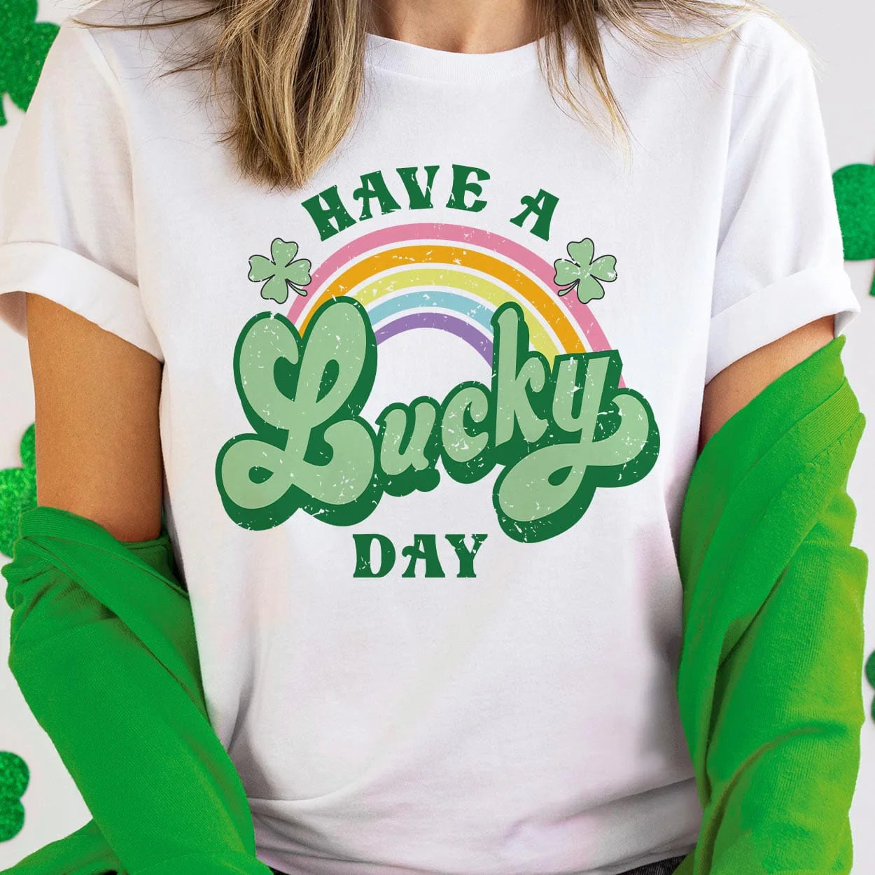 A white short sleeve shirt featuring the words "Have a lucky day" with a rainbow going through the middle with clovers. The word "lucky" is in cursive and a larger font. Item is pictured in a white and green background.