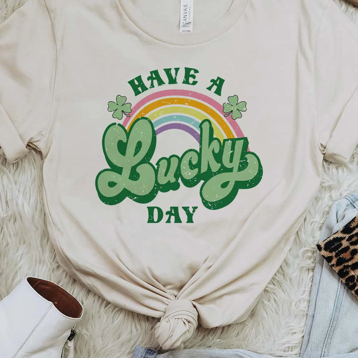 A cream short sleeve shirt featuring the words "Have a lucky day" with a rainbow going through the middle with clovers. The word "lucky" is in cursive and a larger font. Item is pictured in a white background.