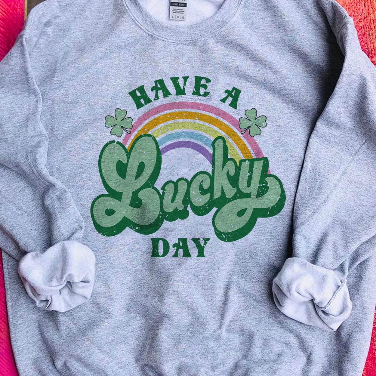 A gray crew neck sweatshirt featuring the words "Have a lucky day" with a rainbow going through the middle with clovers. The word "lucky" is in cursive and a larger font. Item is pictured in a pink background.