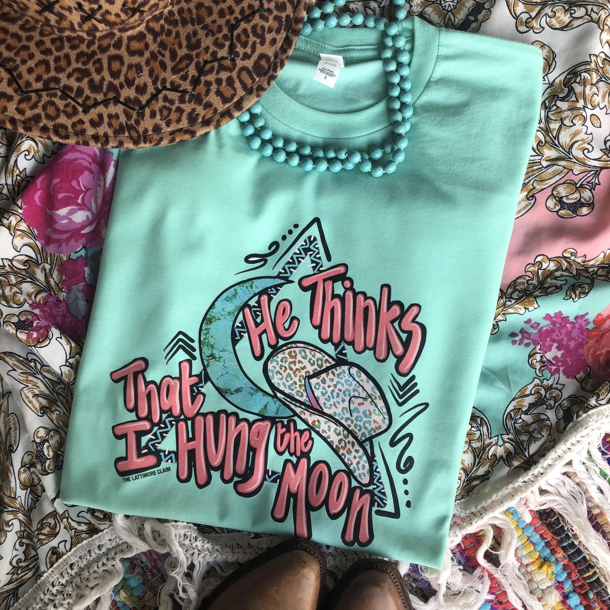 This Bella + Canvas tee in Mint includes a crew neckline, short sleeves, and a cute hand drawn design of a leopard cowboy hat, a turquoise moon, triangle with Aztec trim , and the words "He Thinks That I Hung the Moon" in pink.