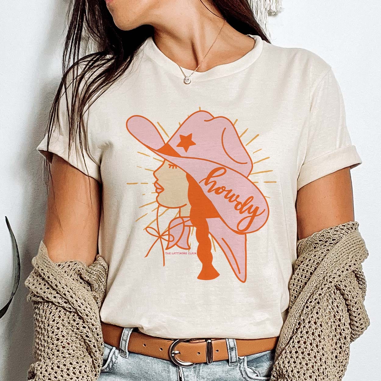 A cream colored short sleeve shirt with folded sleeves featuring a graphic of a cowgirl's side profile with a red braid, pink hat with an orange star and the word "Howdy", a pink bandana, and orange rays coming from the cowgirl. Item is pictured on a plain white background