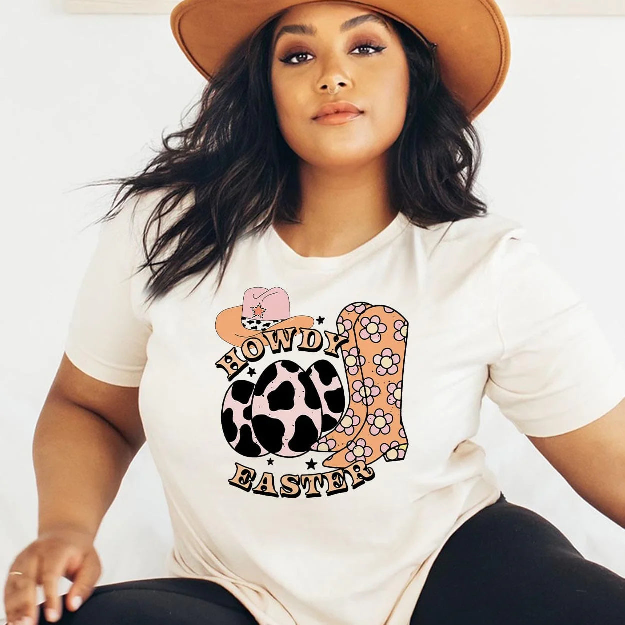 A cream colored short sleeve tee featuring 3 little cow print eggs, a pair of orange floral boots to the right, and a cowboy hat with pink, cow print, and orange above the eggs. The words "Howdy Easter" are curved around the eggs with black little stars. Item is pictured on a plain white background.