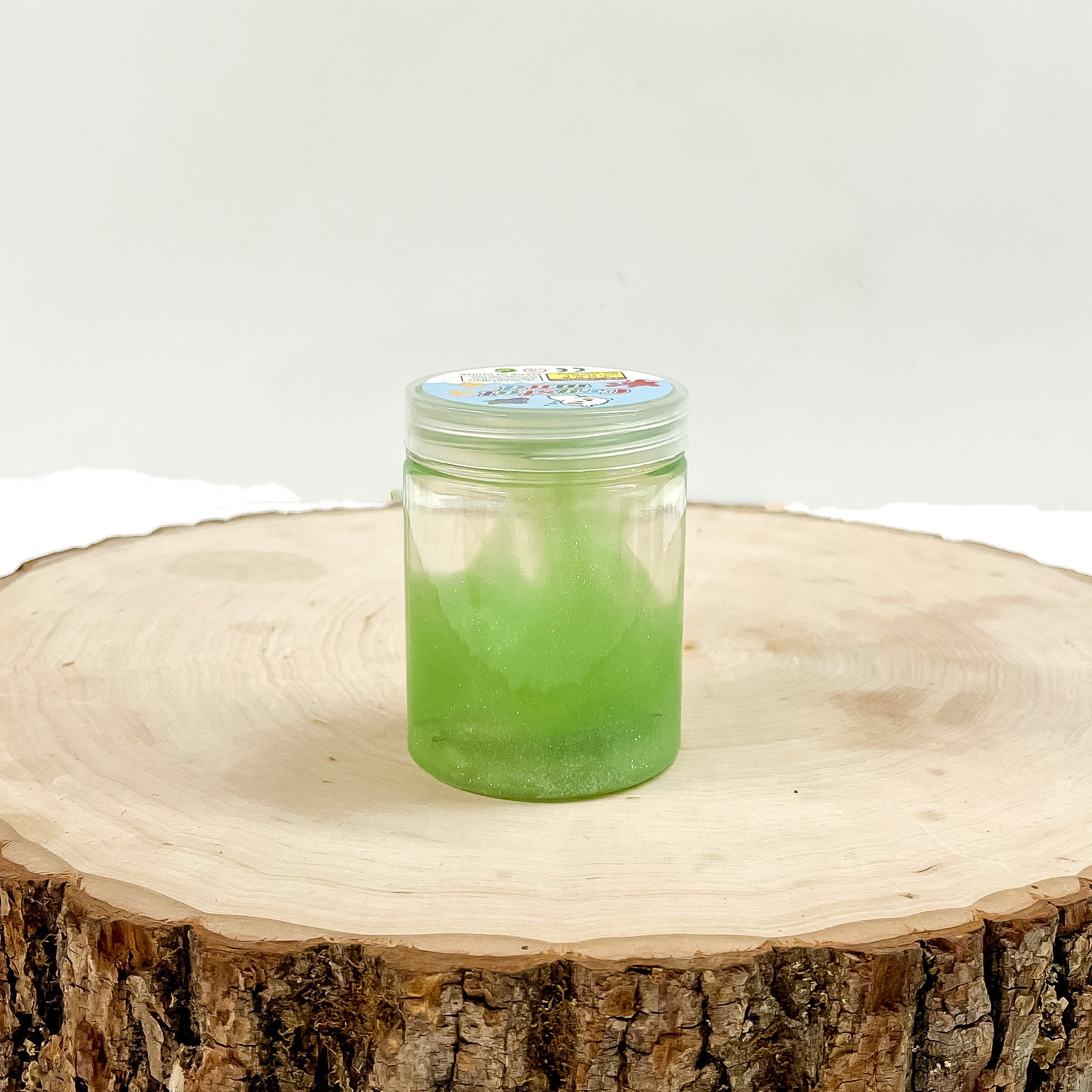 This is a clear slime with green glitter, this container is taken on a slab of  wood and on a white background.