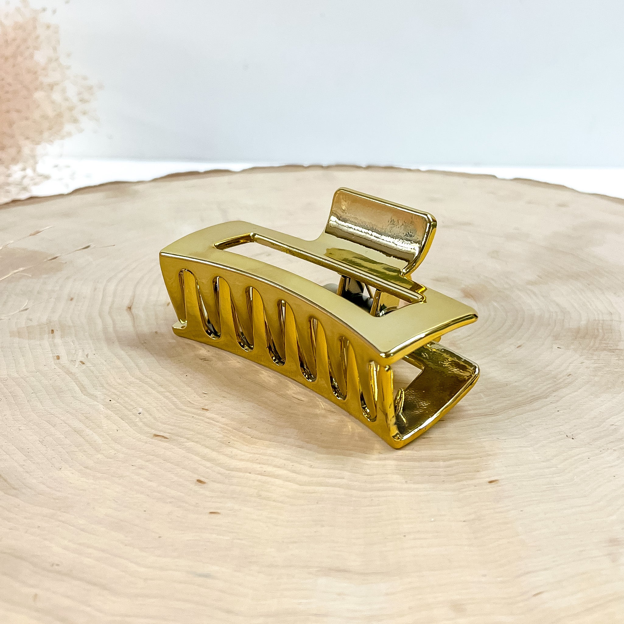 This is a rectangle metallic hair clip in gold, this clip is taken on a  slab of wood and in white background with a plant in the side as decor.
