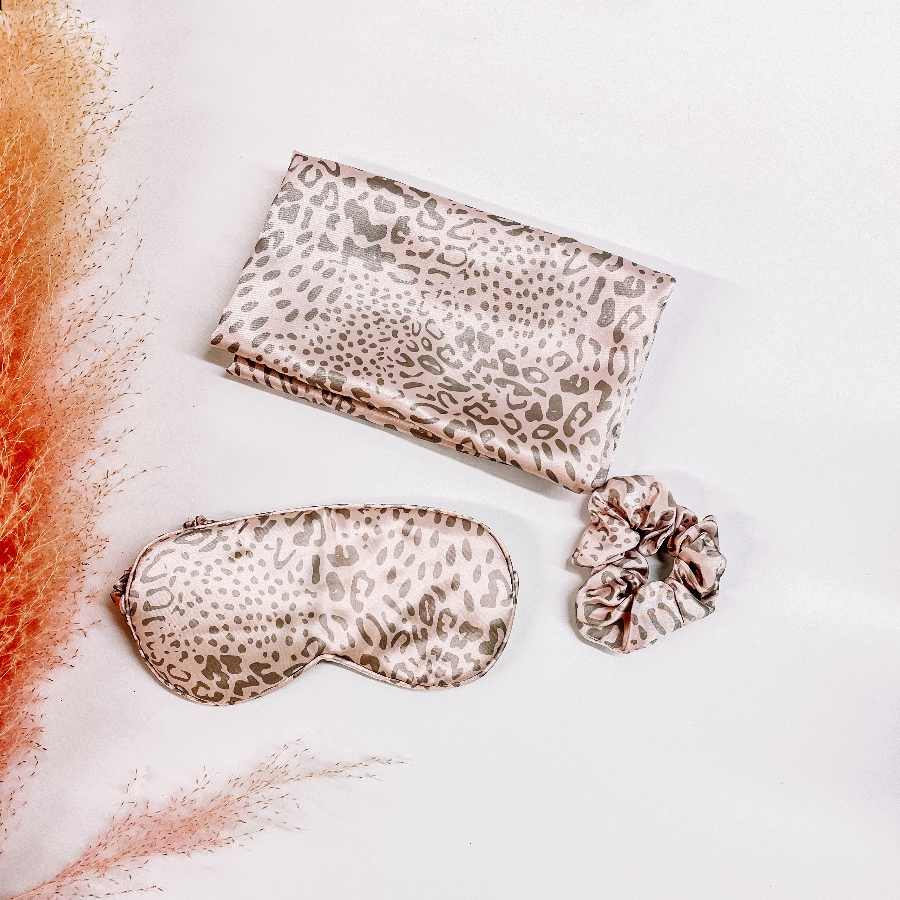There is a satin leopard print pillow case, eye mask, and a scrunchie. The leopard print is in light pink and grey. These items are taken on a white background with a pink plant in the side as decor.