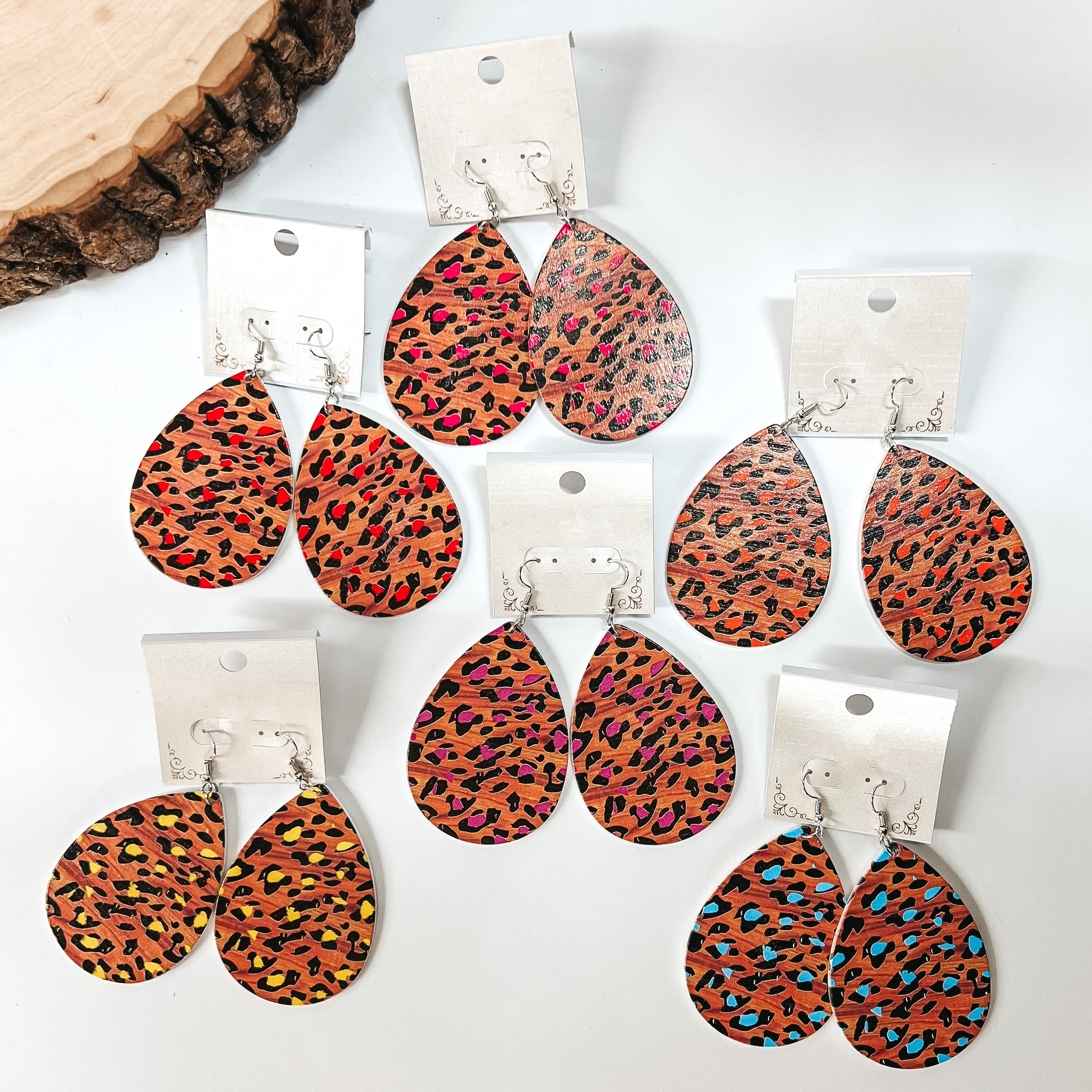 There are six pairs of wooden teardrop earrings in leopard print with different colors. Colors come in pink, purple, red, orange, yellow, and light blue. All earrings are placed in a white earrings holder. These earrings are taken on a white background with a slab of wood in the back as decor.