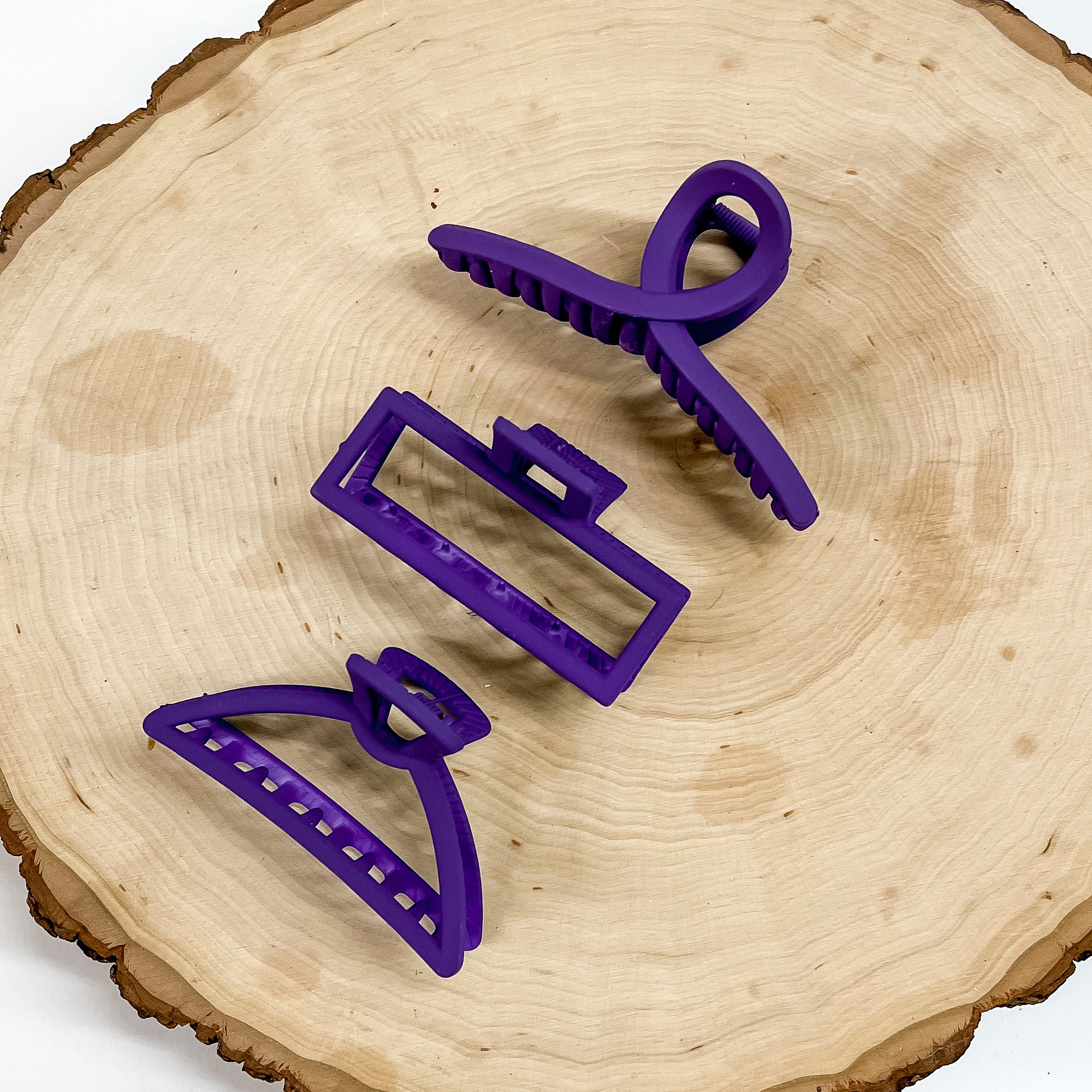 Pictured on a piece of wood are 3 matte purple hair clips. The first is a loop clip, a rectangle clip, and a triangle clip. 