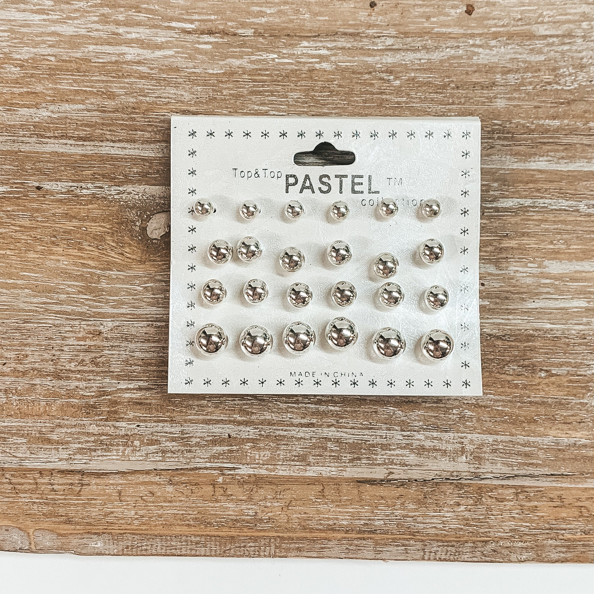 This is a pack of twelve silver circle stud earrings in different sizes. These  earrings are placed on a white earring card holder and laying on a wooden slate.