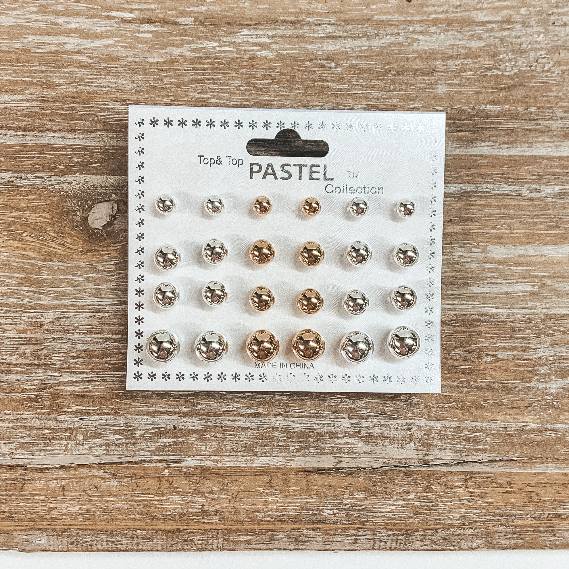 This is a pack of twelve circle stud earrings in different sizes and colors such as gold and silver. These earrings are placed on a white earring card holder and laying on a wooden slate.