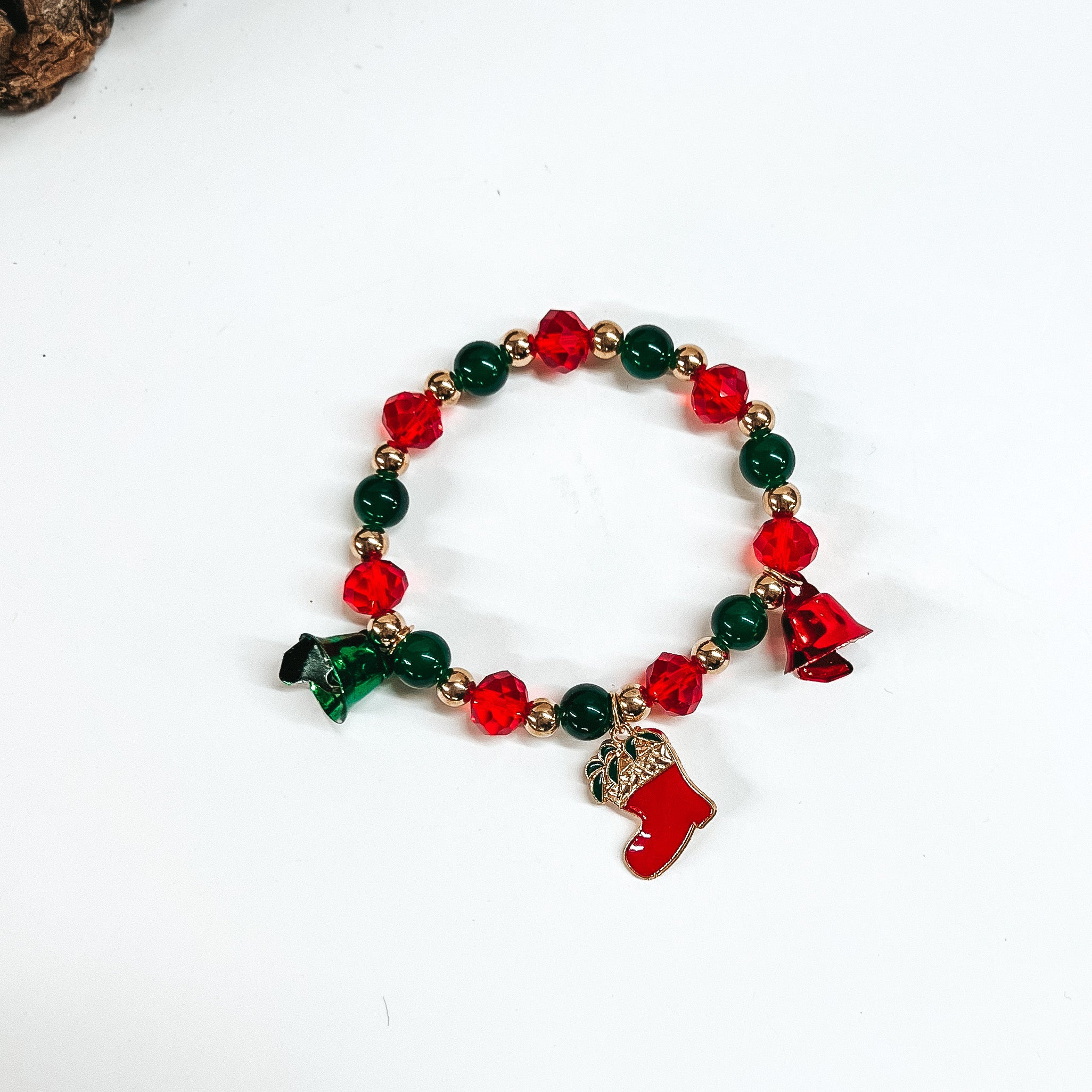 This is a beaded bracelet with three christmas charms such as two bells in red and green, and a gold and red stocking charm. This beaded bracelet has red crystal beads, gold circle beads, and green circle beads. This bracelet is laying on a white background with a slab of wood in the back as decor.