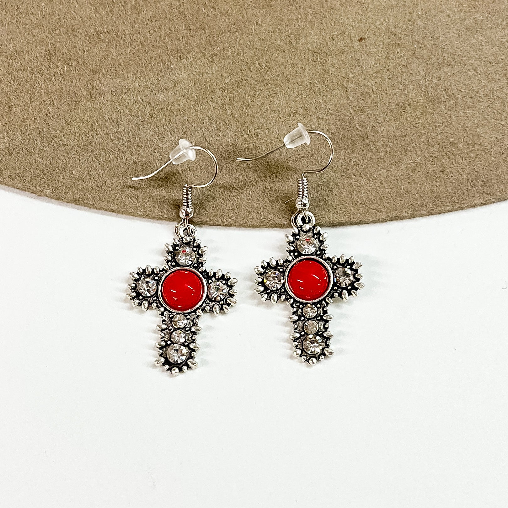 This is a pair of silver detailed cross earrings with small clear crystals  and a red circle stone in the center.  These earrings are laying on a brown felt hat brim and on a white background.