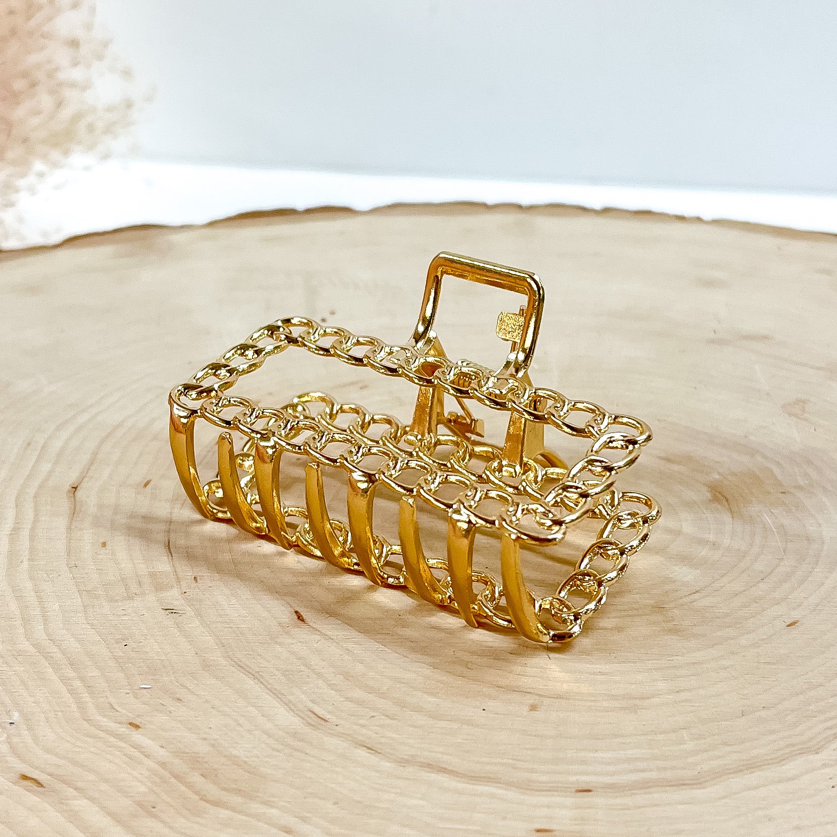 This is a small rectangle shaped hair clip with chain texture in gold. This hair  clip is laying on a slab of wood with a beige plant in the side as decor.