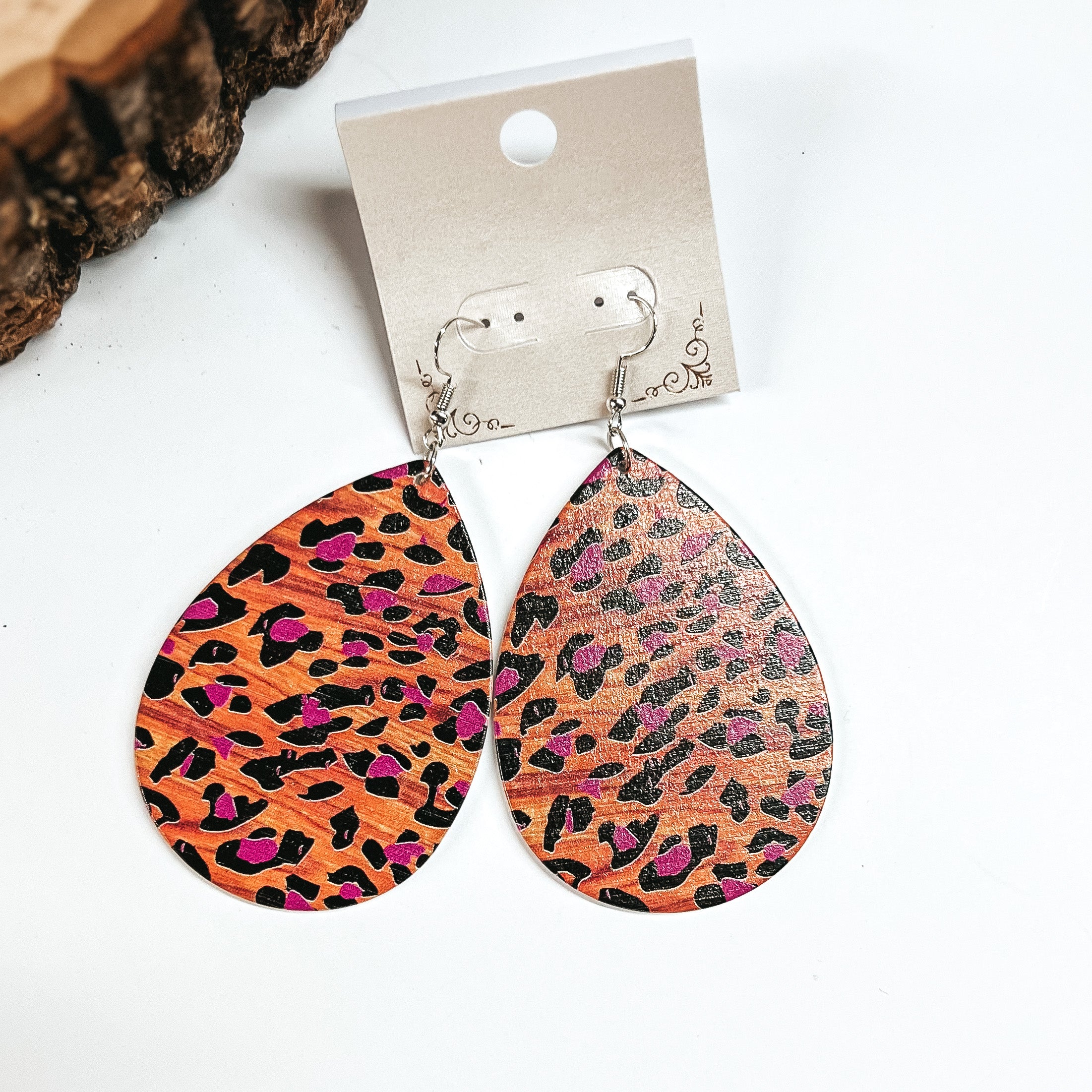 These are brown wooden teardrop earrings in leopard print and purple detailing. These earrings are placed on a white earring card holder, they are laying on a  white background with a slab of wood in the back as decor.