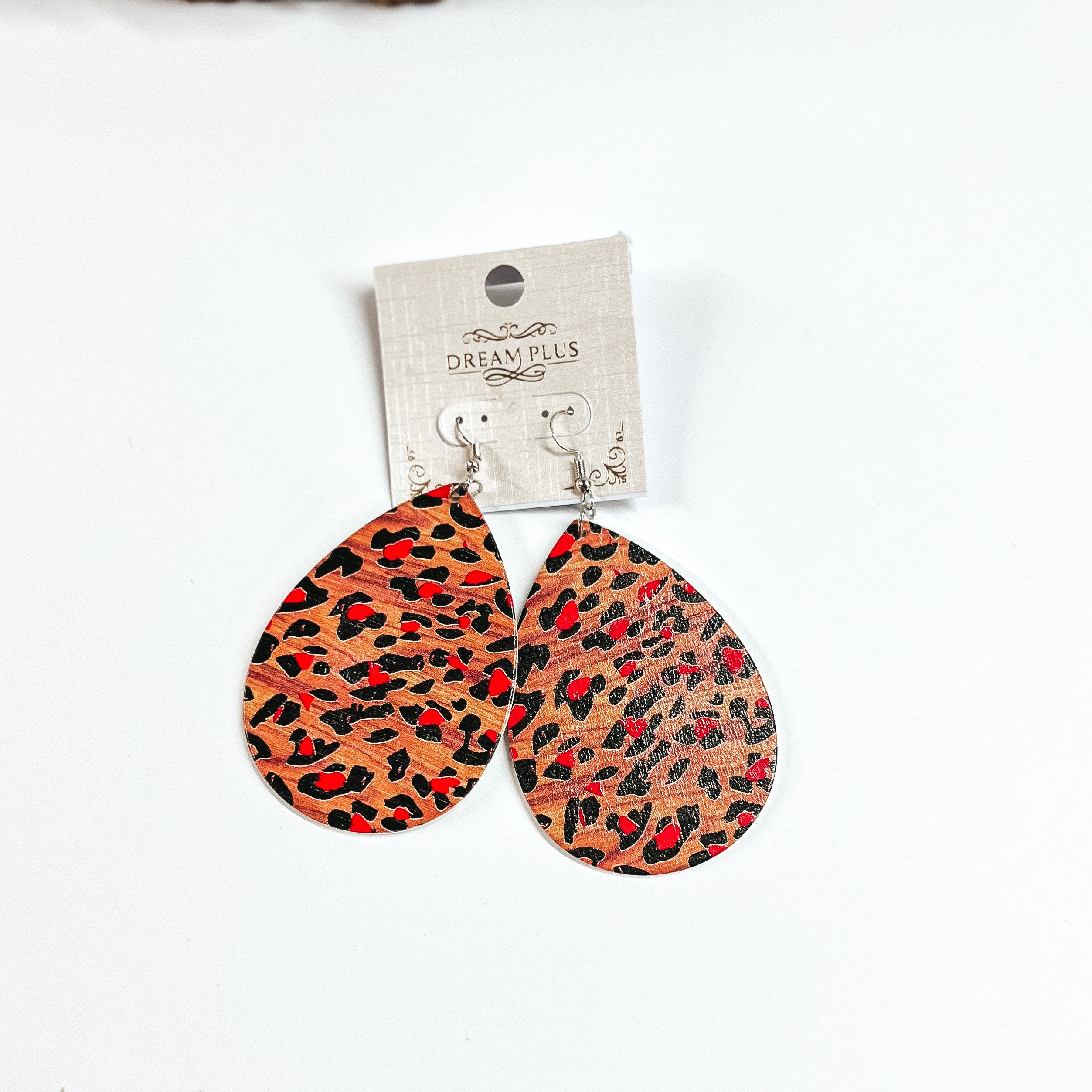 These are brown wooden teardrop earrings in leopard print and red detailing. These earrings are placed on a white earring card holder, they are laying on a  white background with a slab of wood in the back as decor.