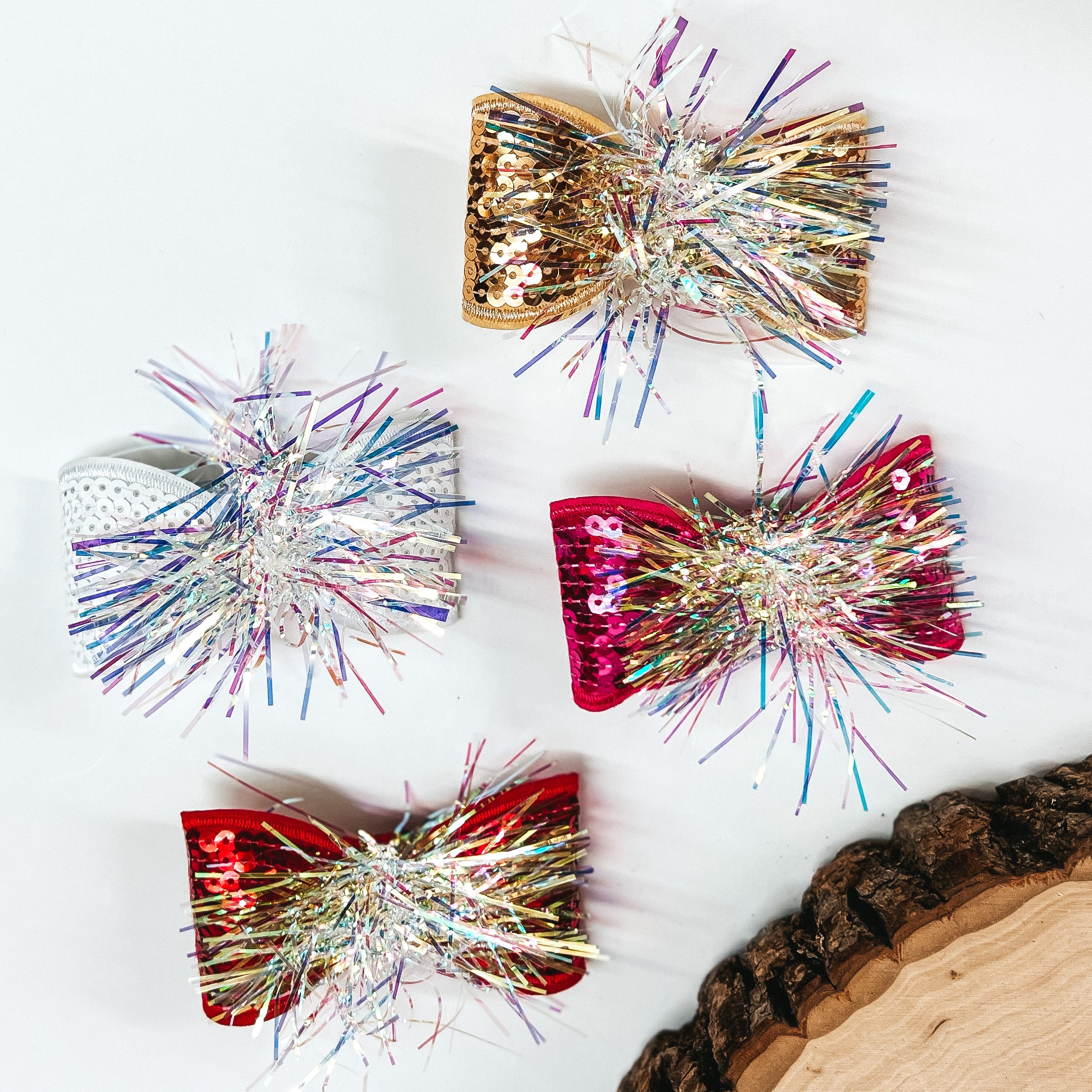 There are four sequins bows with a tinsel center. From top to bottom; gold, white, hot pink, and red. These bows are laying on a white background with a slab of wood in the corner as decor.