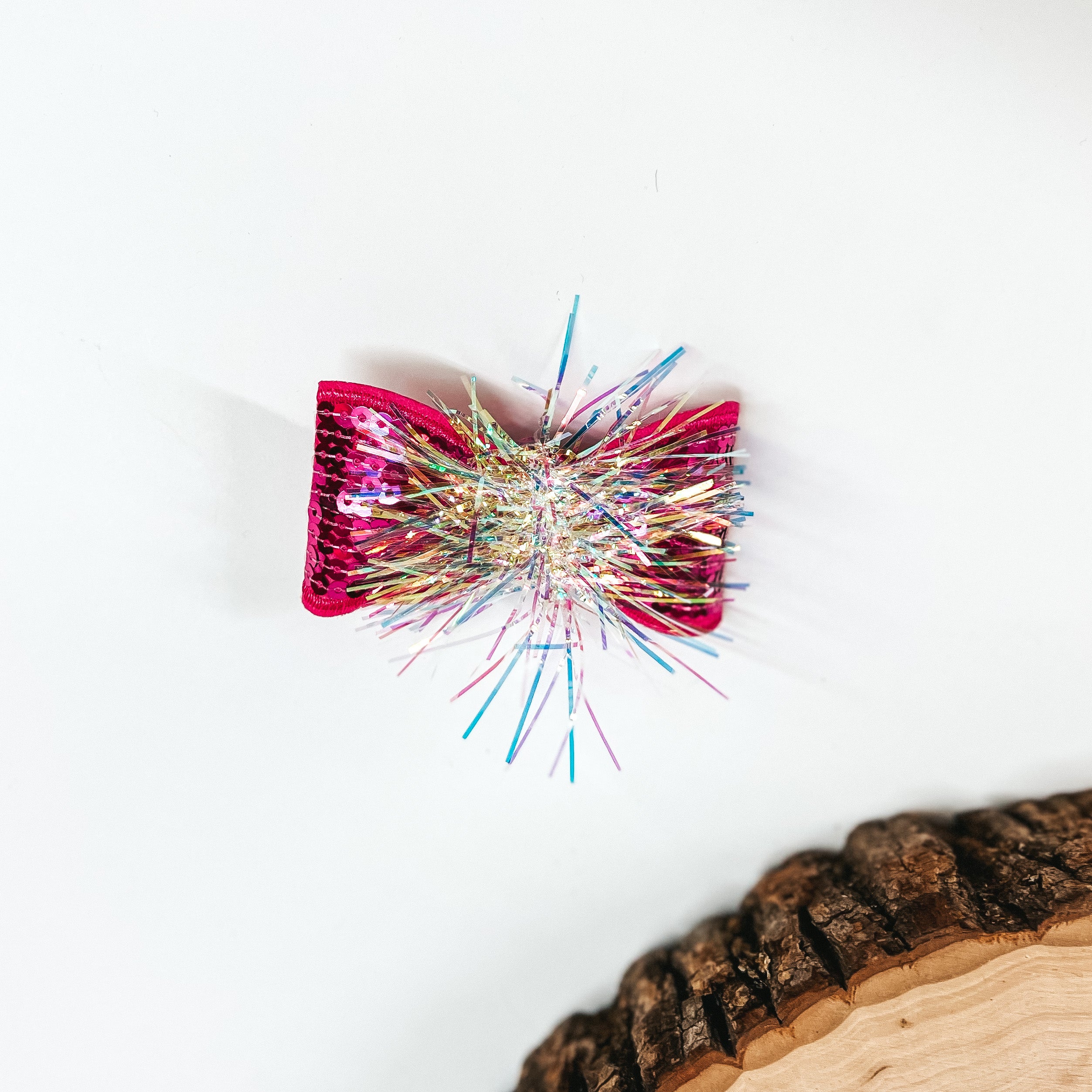 This is a small hot pink sequin bow with a tinsel center, this bow is taken on a  white background with a slab of wood in the corner as decor.